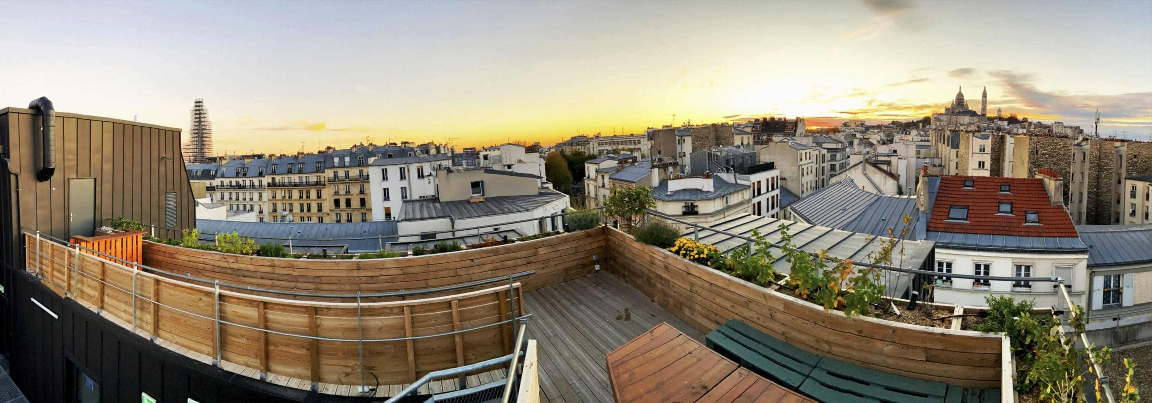 A panoramic view of a Paris rooftop at sunset, showcasing the cityscape with buildings, rooftops, and the outline of Montmartre in the distance.