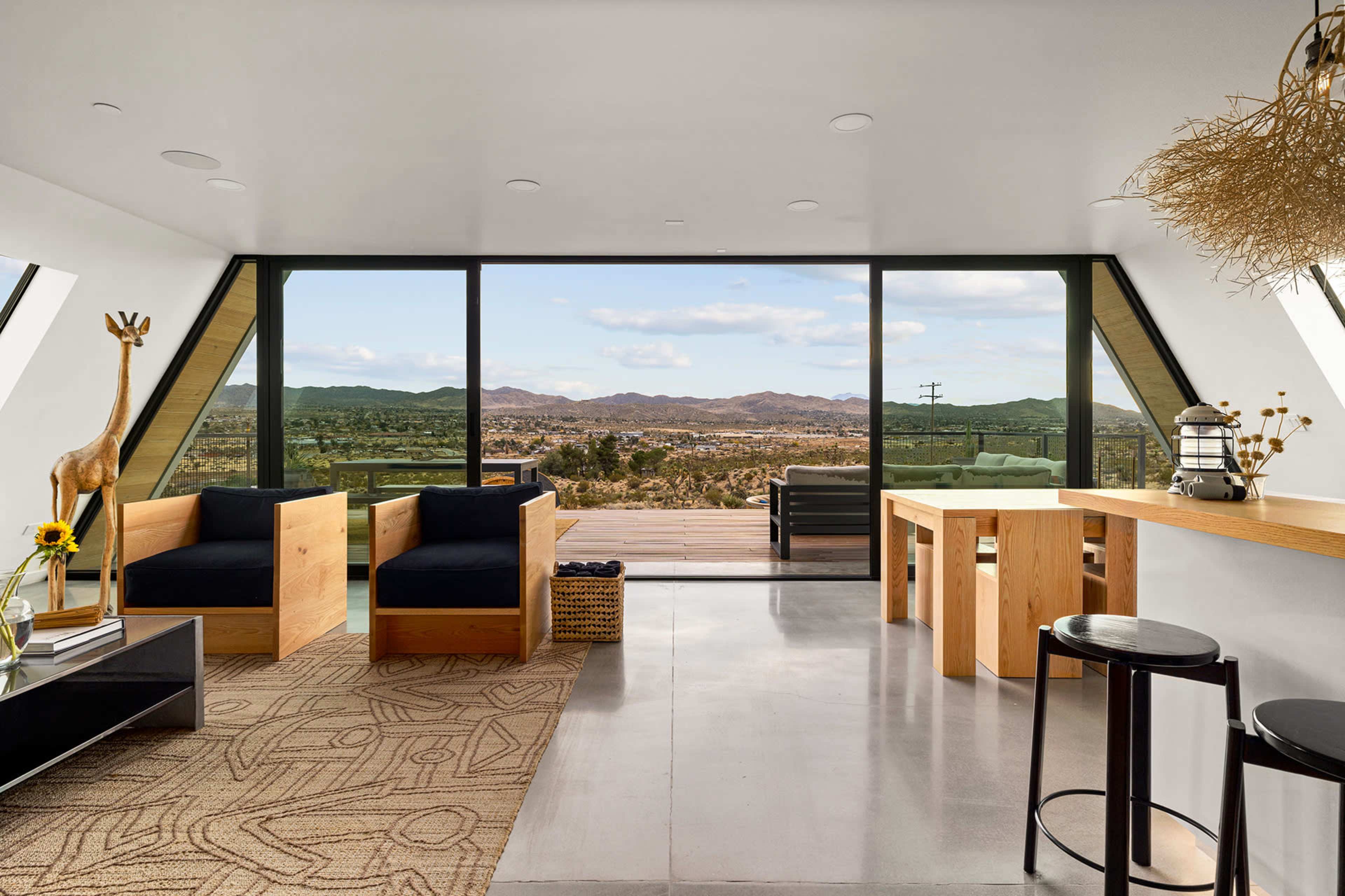 The image shows a modern interior space with large windows that provide a view of a desert landscape, featuring wooden furniture and minimalist decor.
