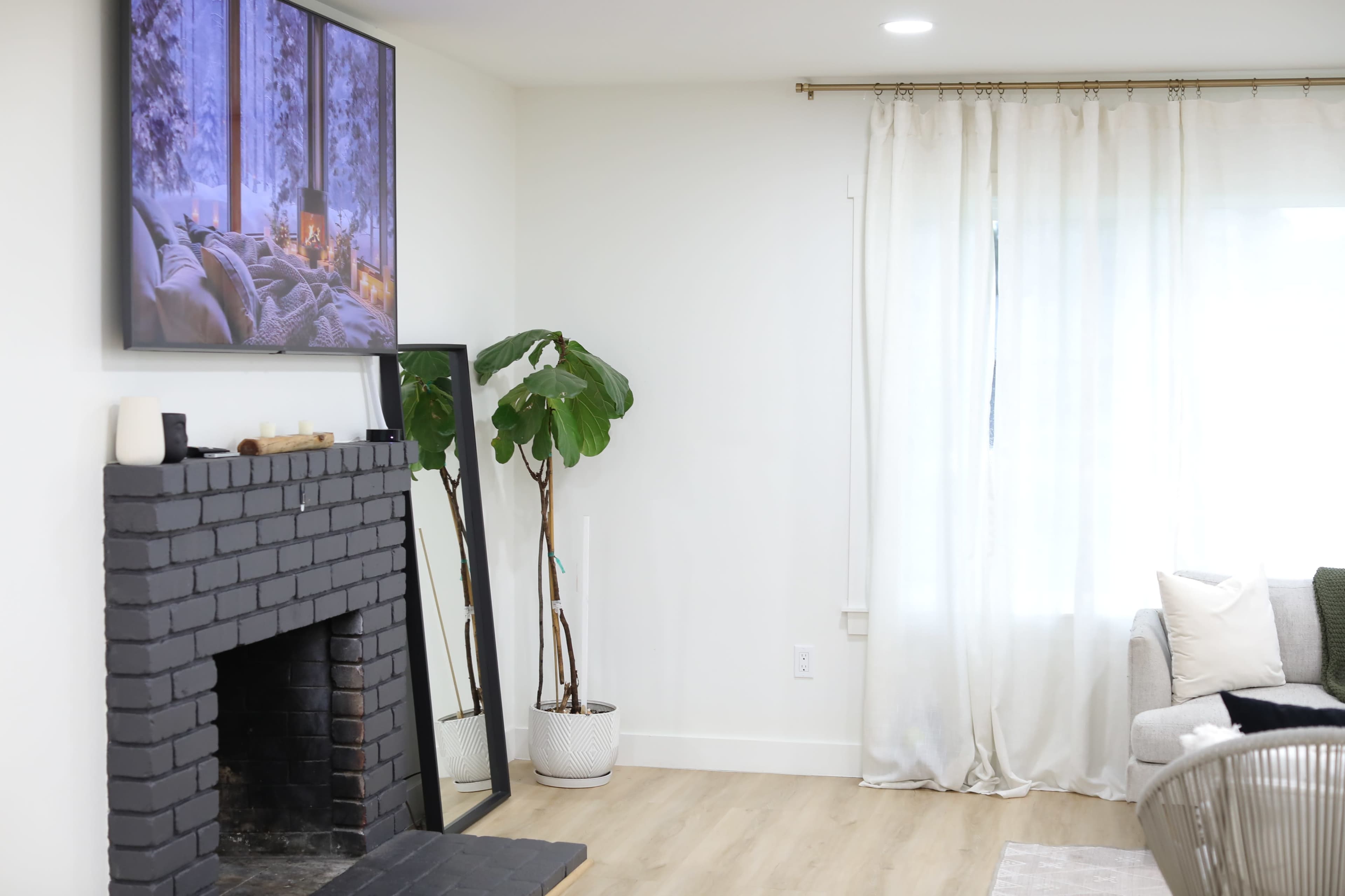 A modern living room features a gray brick fireplace, a flat-screen TV mounted above it, and a large window with light-colored curtains.