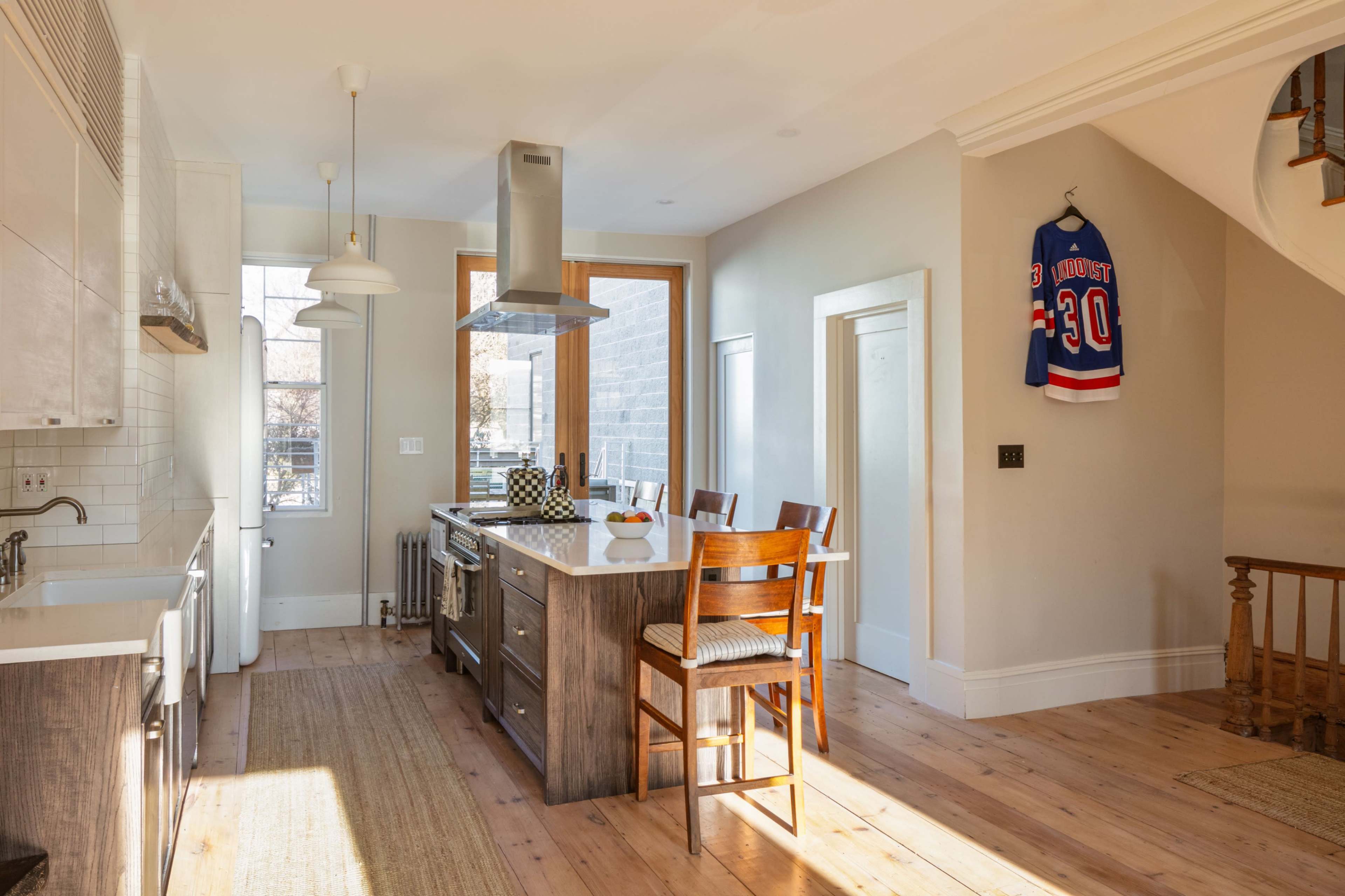 The image shows a modern kitchen with wooden flooring, a central island with bar stools, and a hockey jersey hanging on the wall.