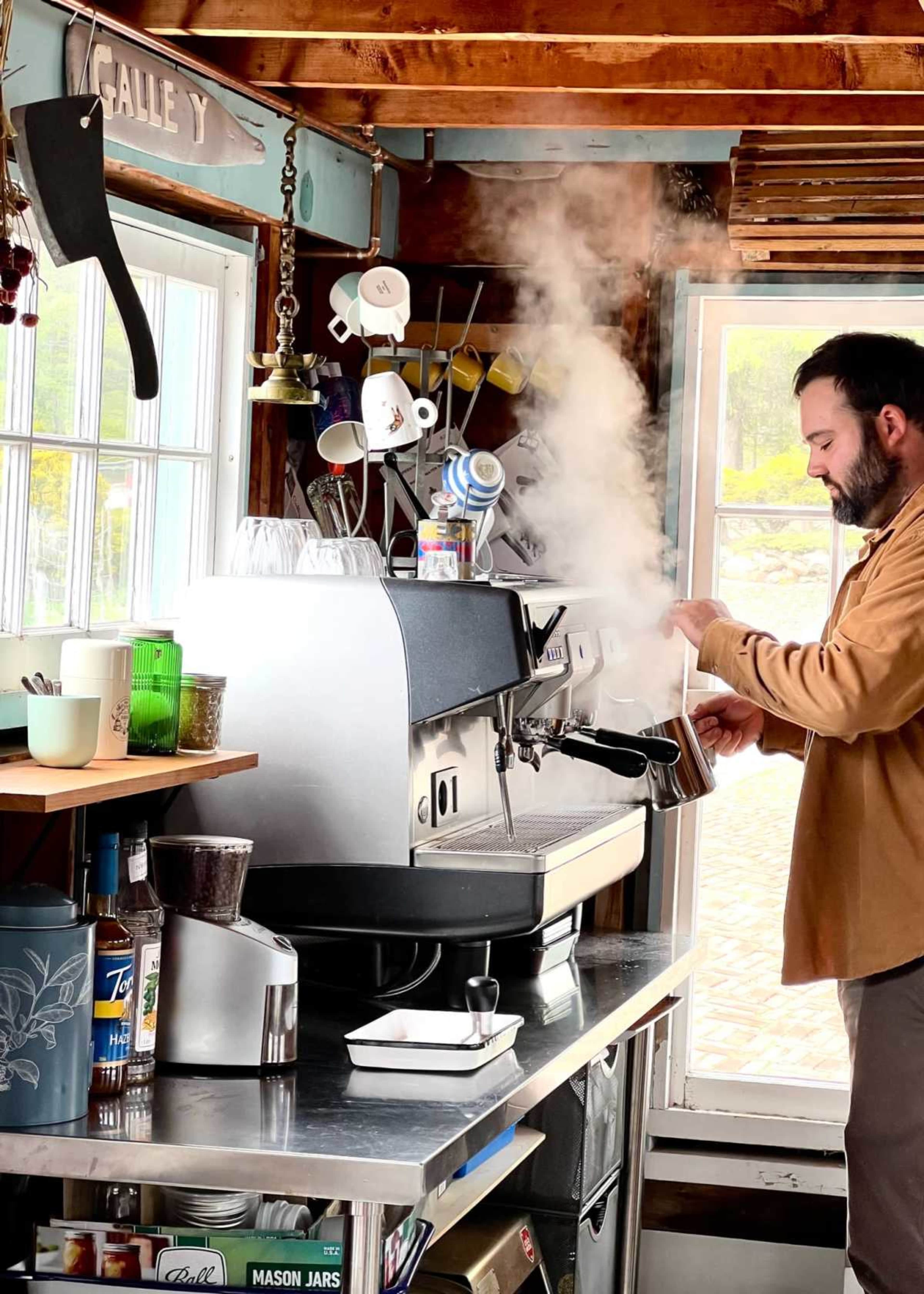 A barista operates an espresso machine in a rustic cafe, with steam rising amidst kitchen utensils and glassware.