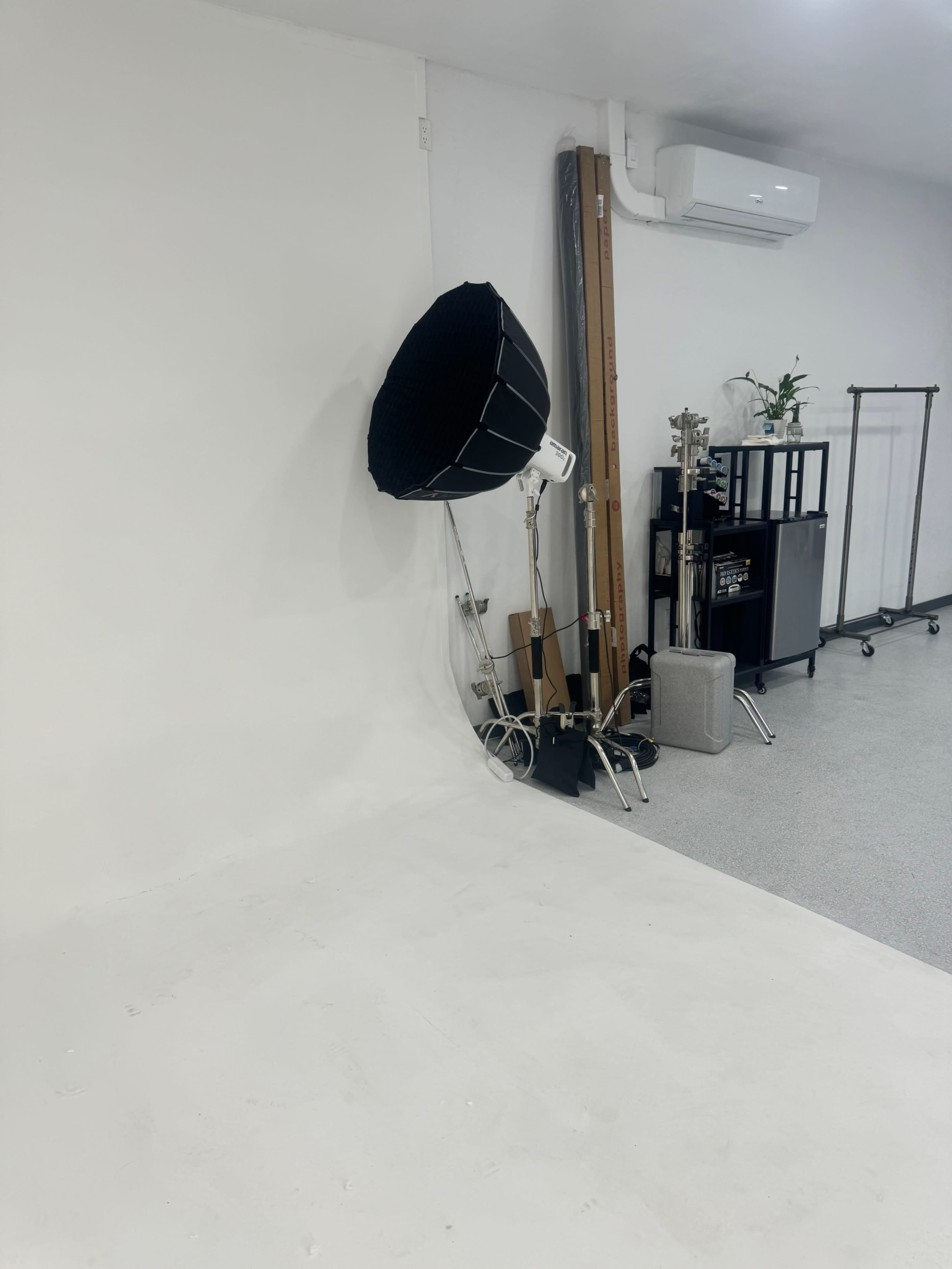 The image shows a photography studio corner featuring a large softbox light on a stand, against a plain white wall, with various studio equipment and a small plant on a shelf nearby.