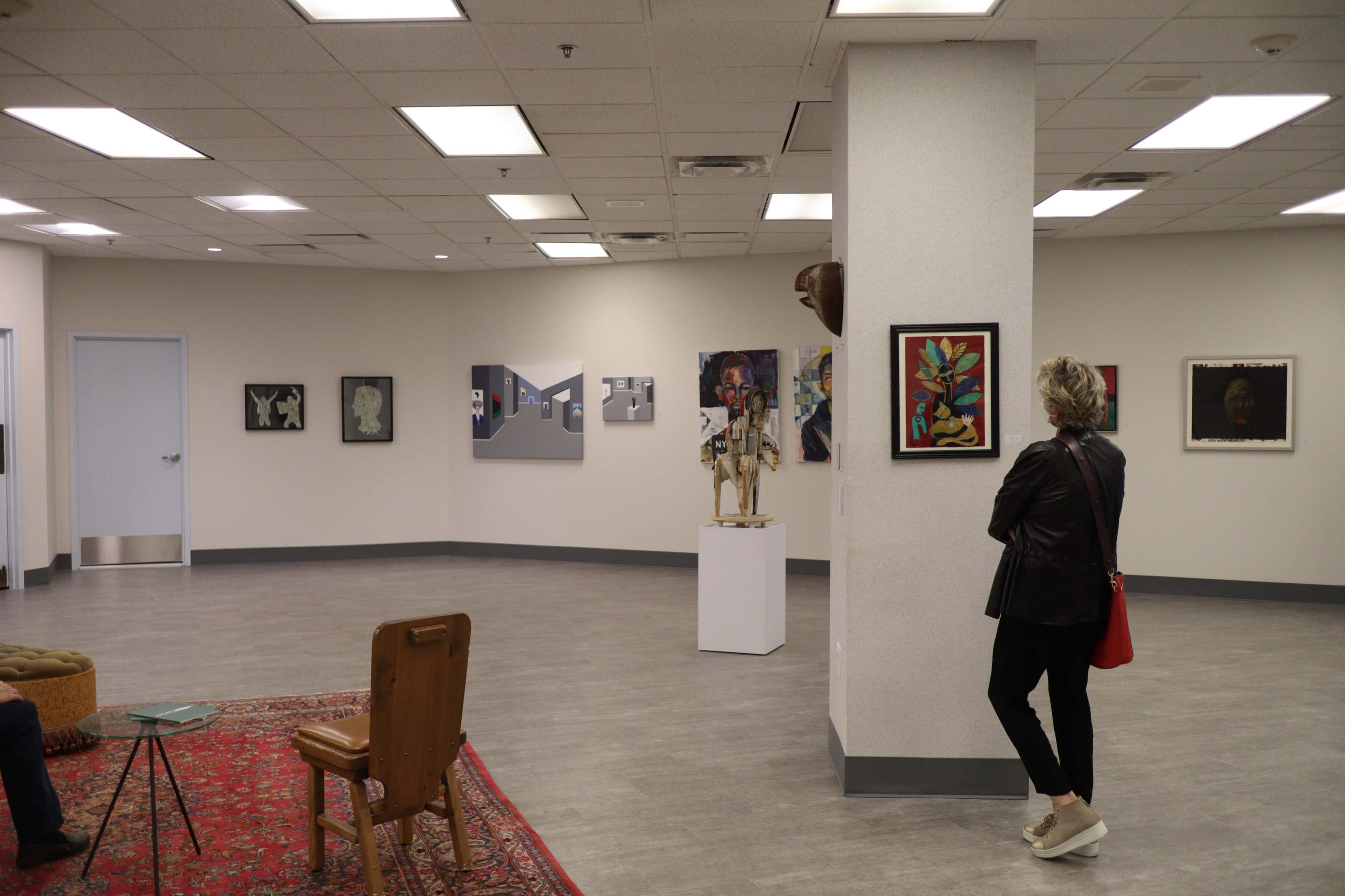 A contemporary art gallery with various artworks displayed on the walls, a wooden chair, and a red rug on the floor.