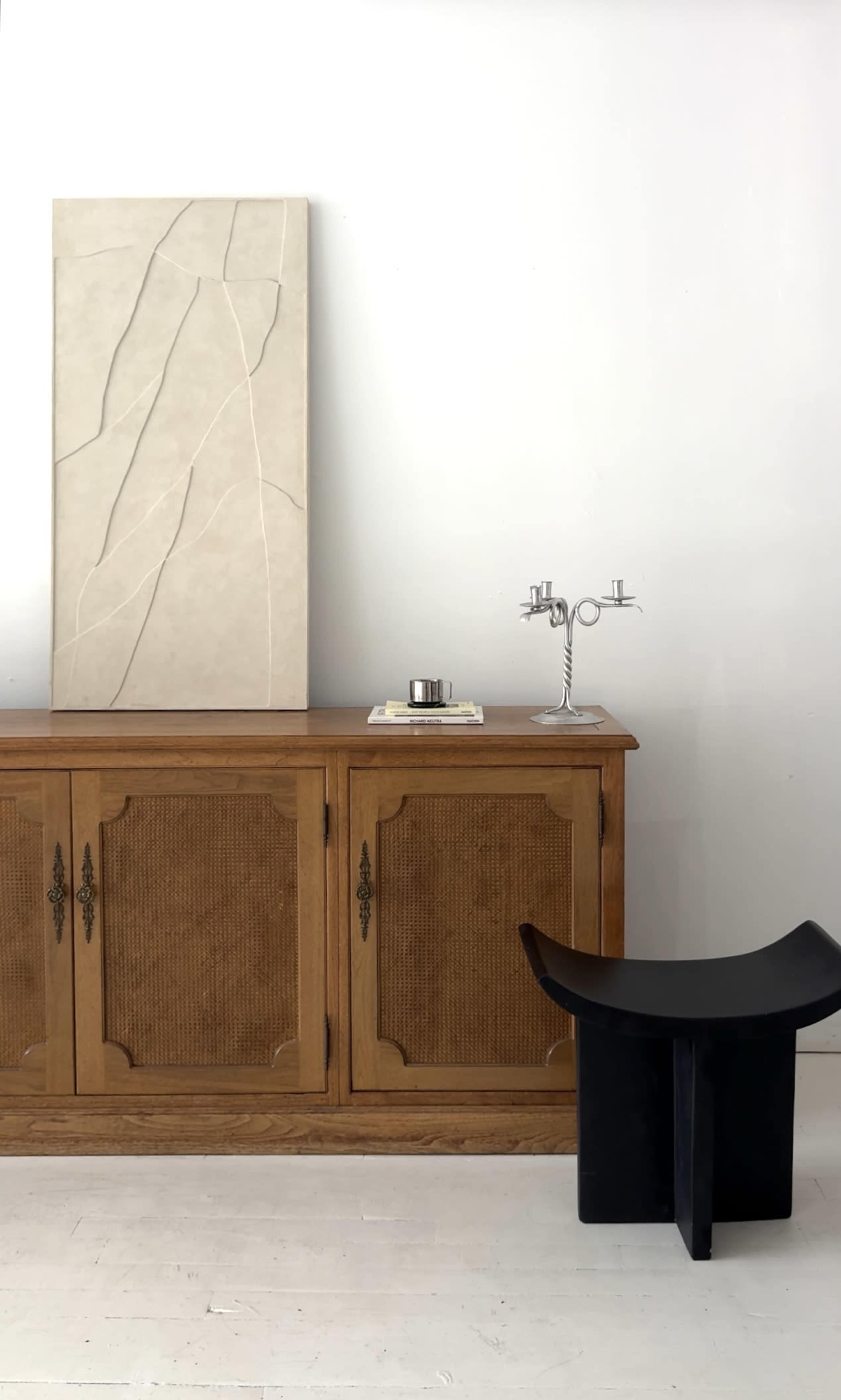 A wooden sideboard with an abstract wall art piece above it, a small tray with a candle, and a black stool are arranged in a minimalist interior.