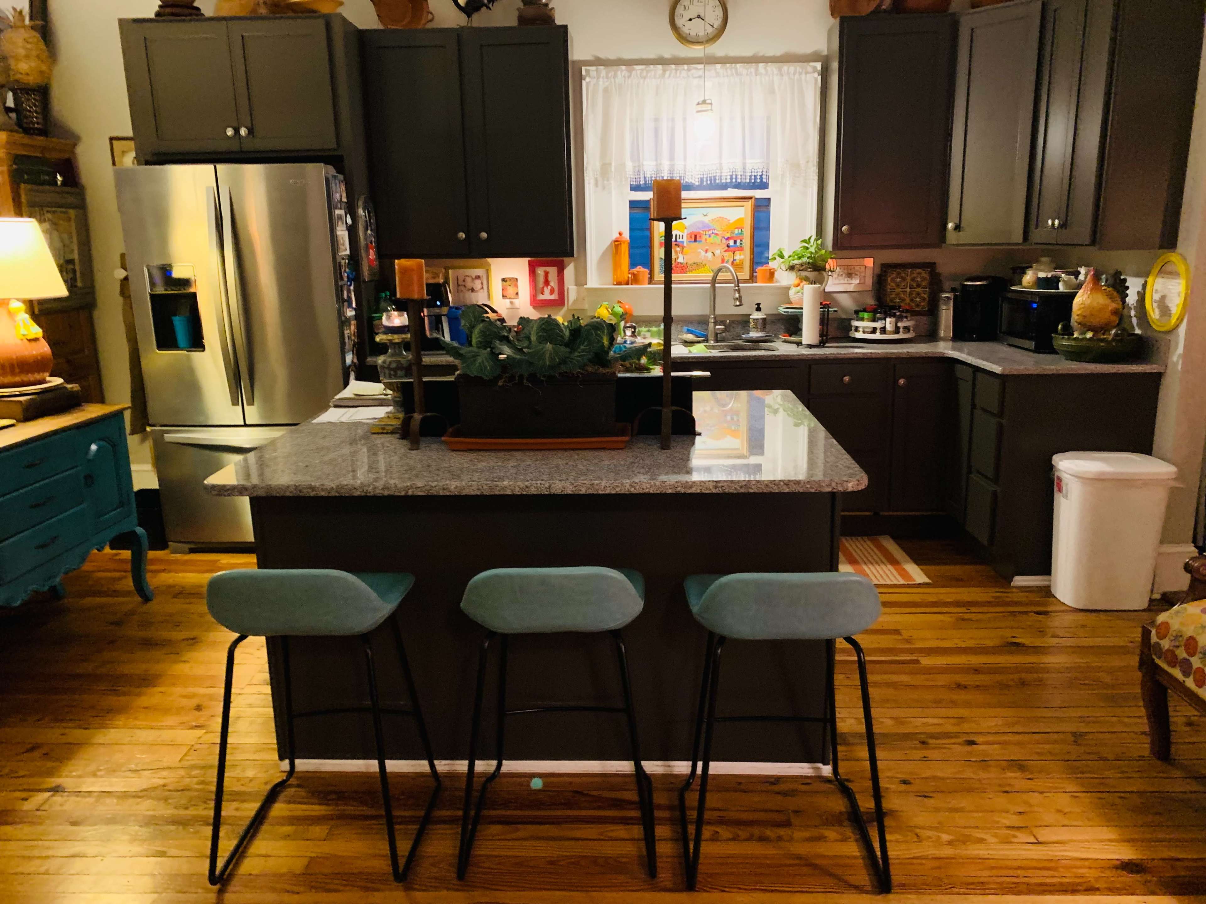 A modern kitchen with dark cabinetry, a central island with three stools, and various appliances and decorations.