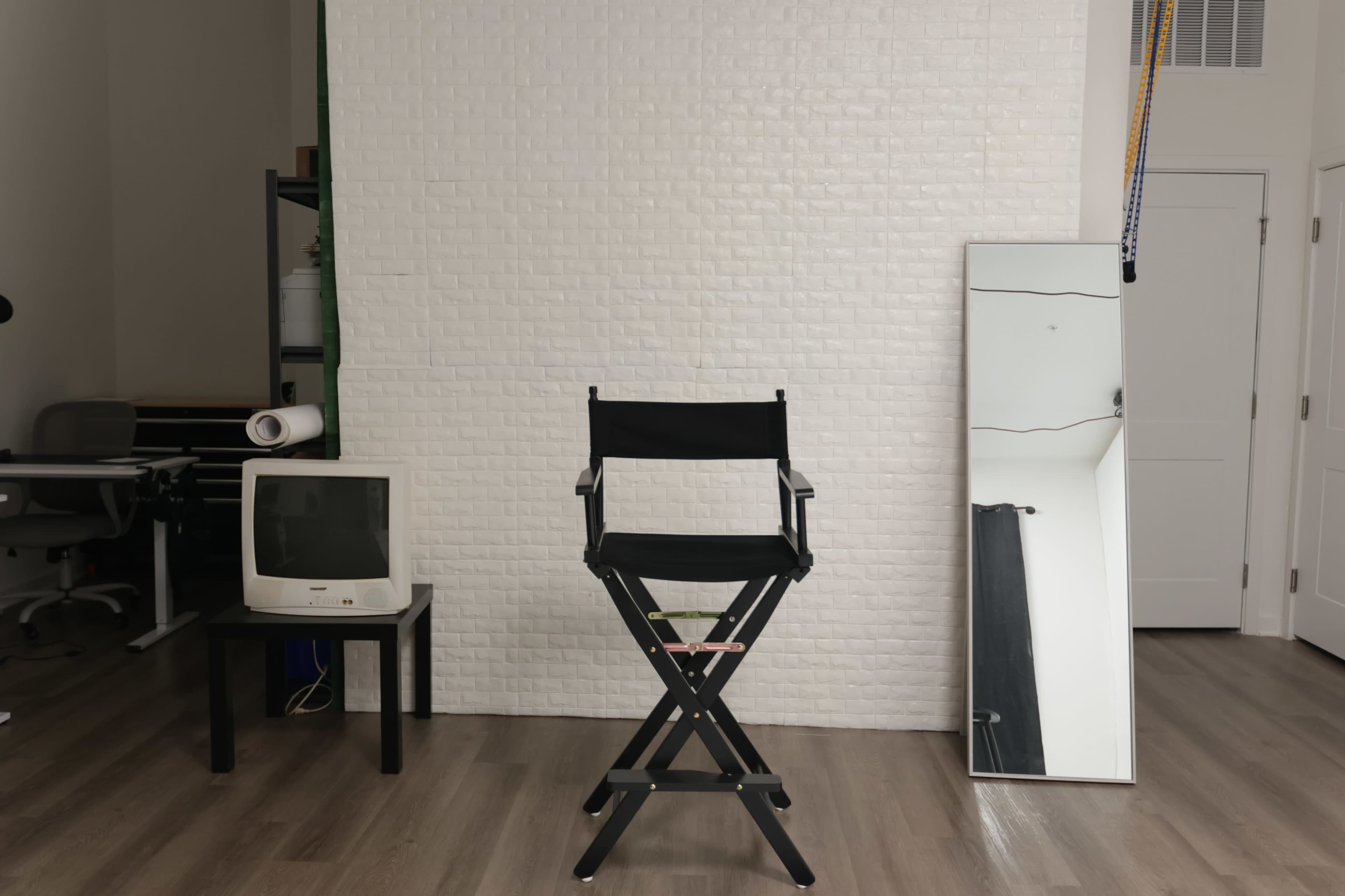 The image shows a minimalist studio setup with a director's chair facing a white brick wall, accompanied by a small CRT television and a large mirror to the side.