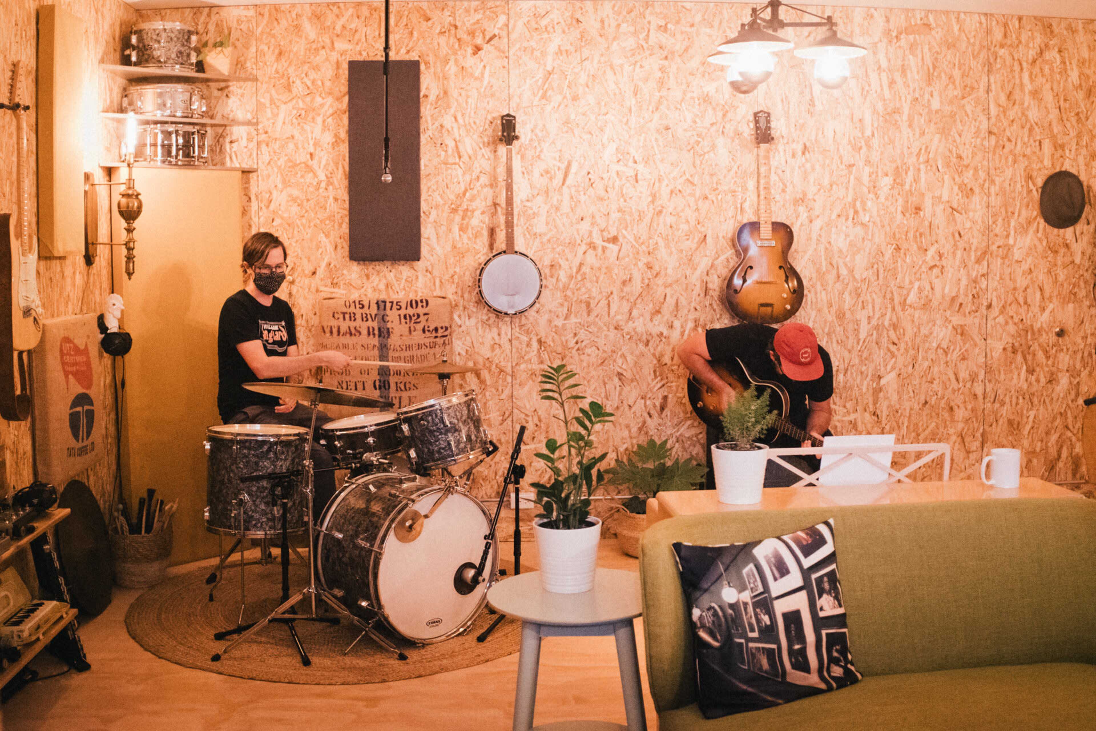 Two musicians play drums and guitar in a cozy room decorated with wooden panels and musical instruments.