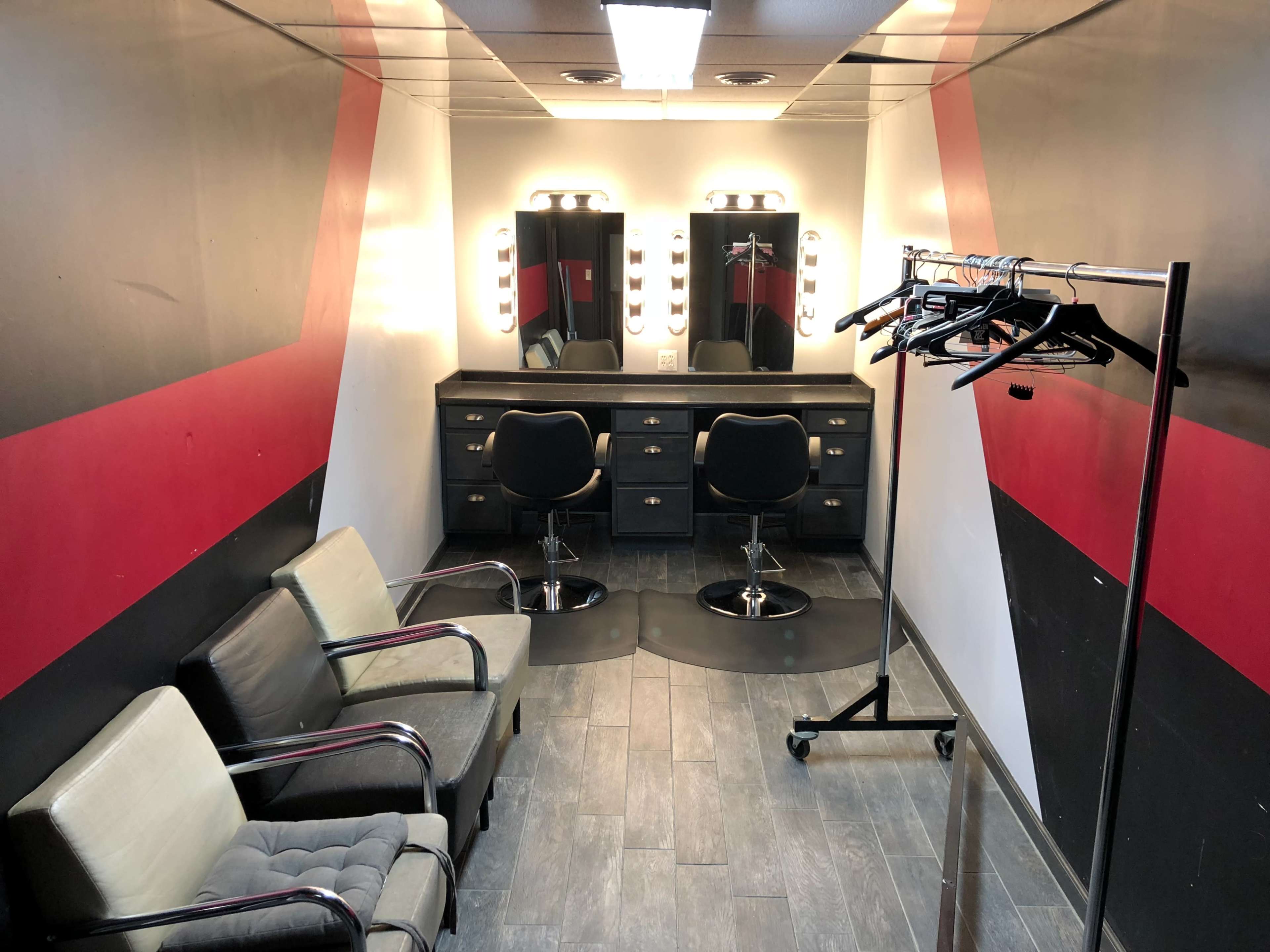The image shows a makeup or dressing room with two illuminated mirrors, black chairs, and a clothing rack lined with hangers.