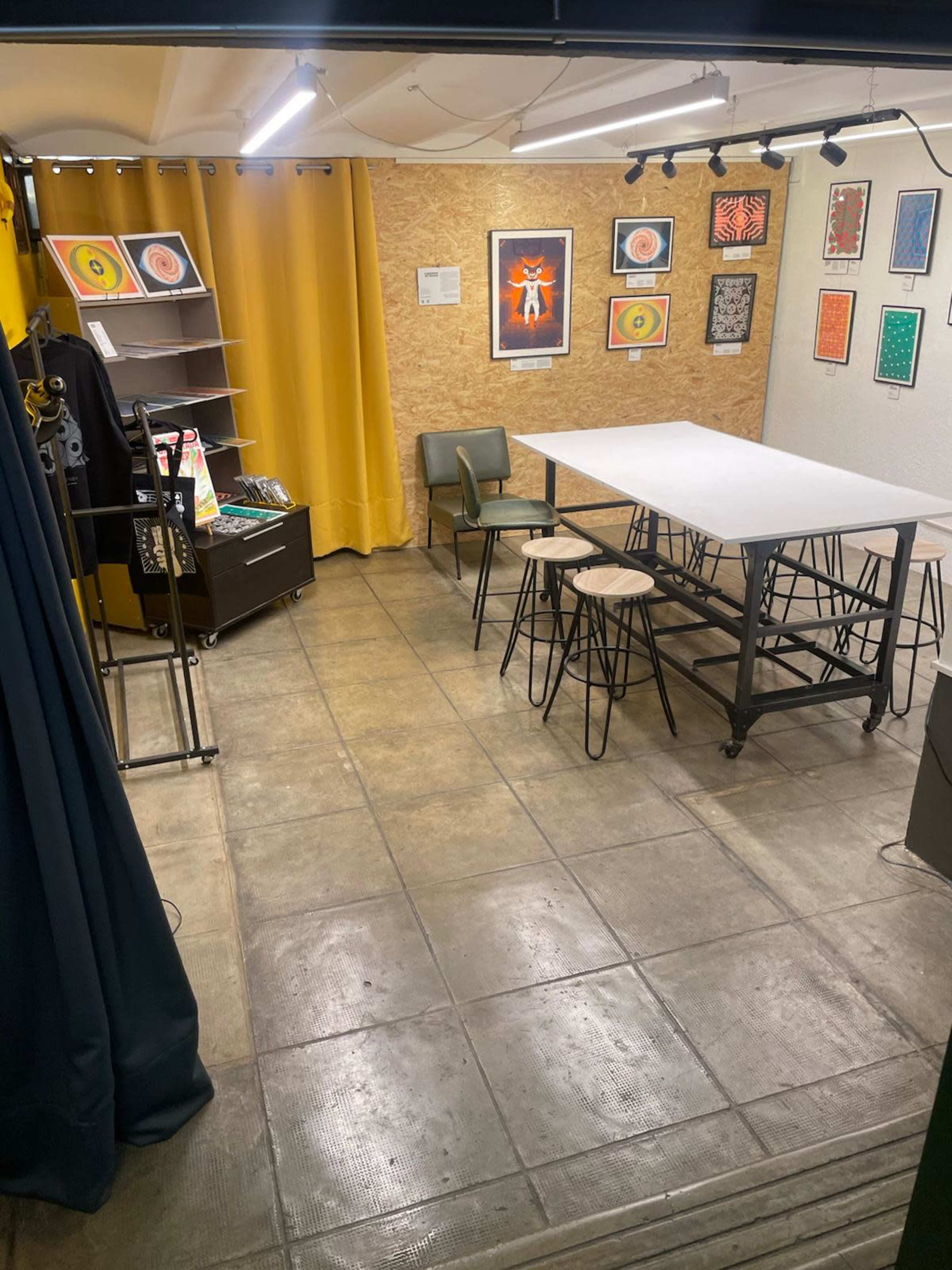 The image shows a spacious, well-lit room with a large white table, several chairs, artwork on the walls, and yellow curtains.