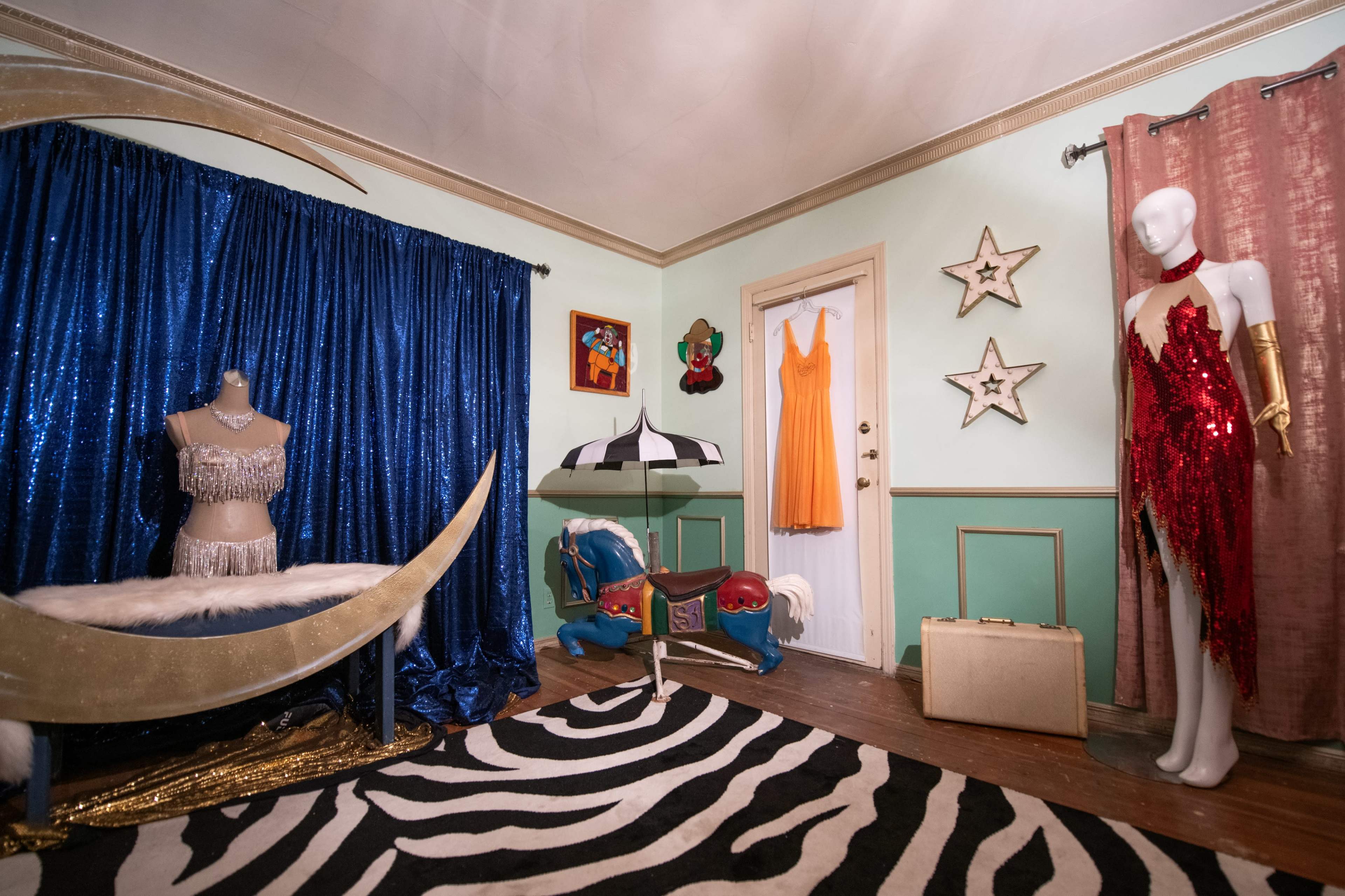 The room features a variety of colorful costumes and decor, including a sequined dress on a mannequin, a blue backdrop, and a whimsical horse-shaped rocking toy.