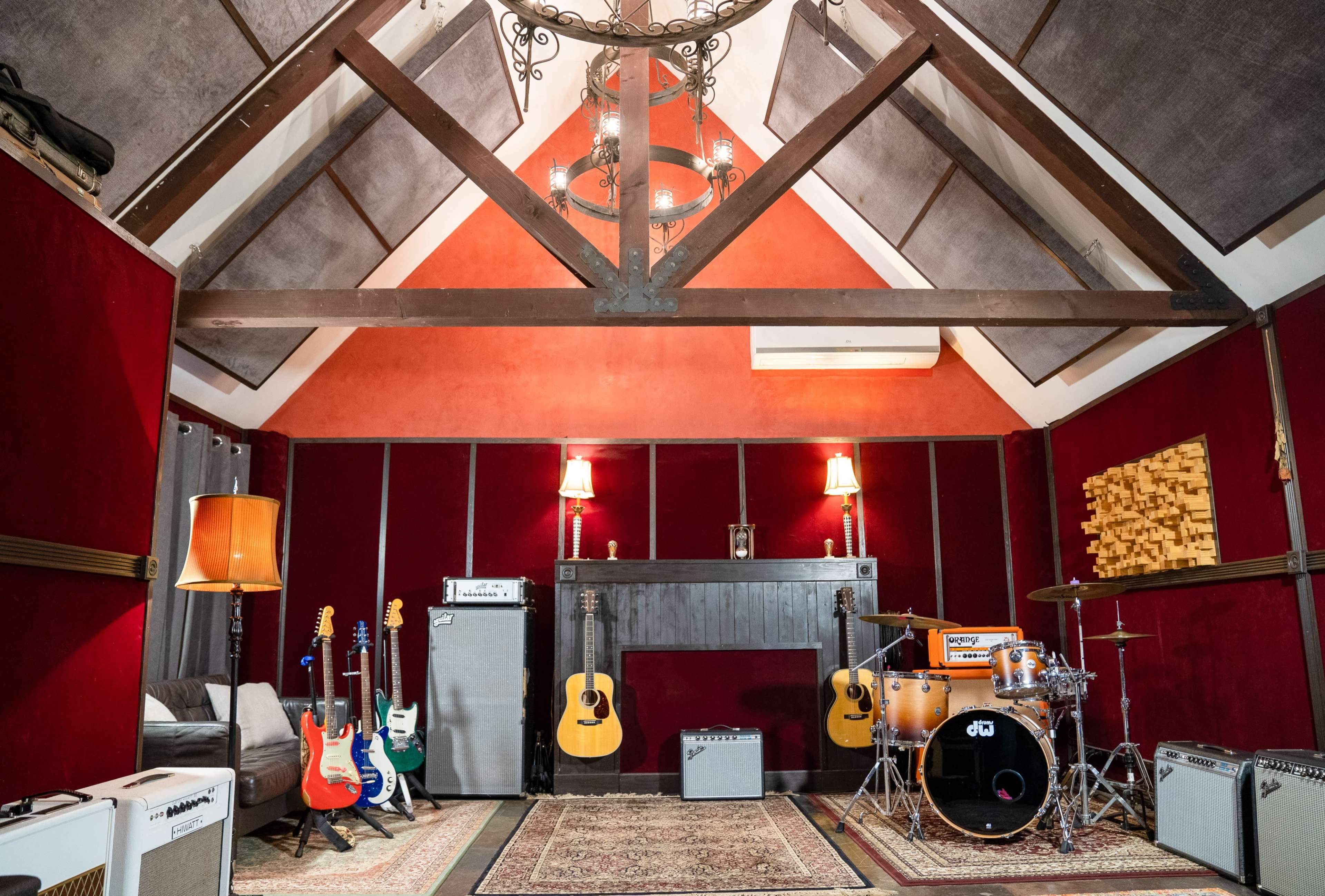 A music recording studio with various guitars, amplifiers, and a drum set arranged within a room featuring wood beams and red-paneled walls.