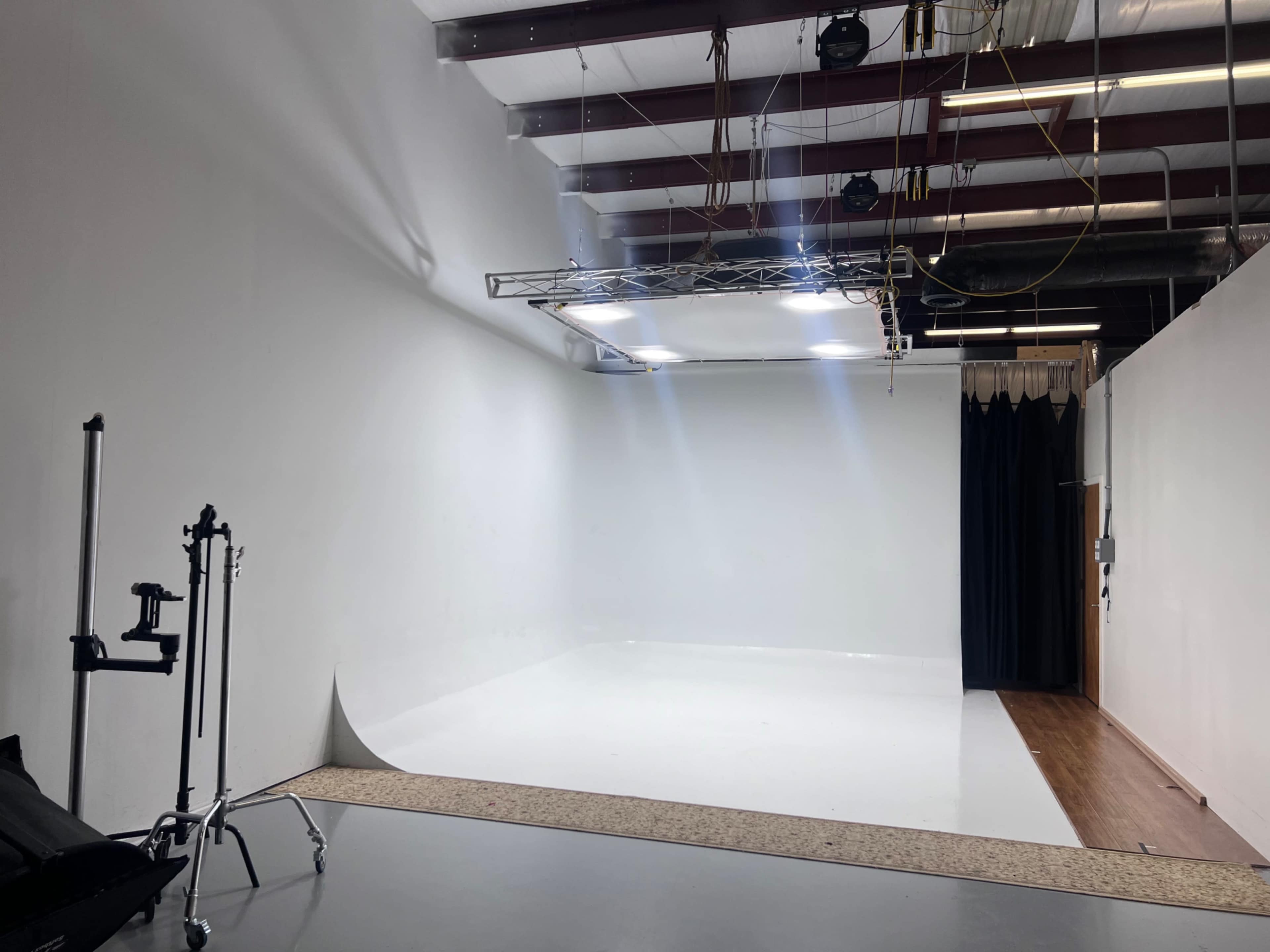 A photo studio features a large white backdrop, bright lights mounted on a truss, and black curtains along one side.
