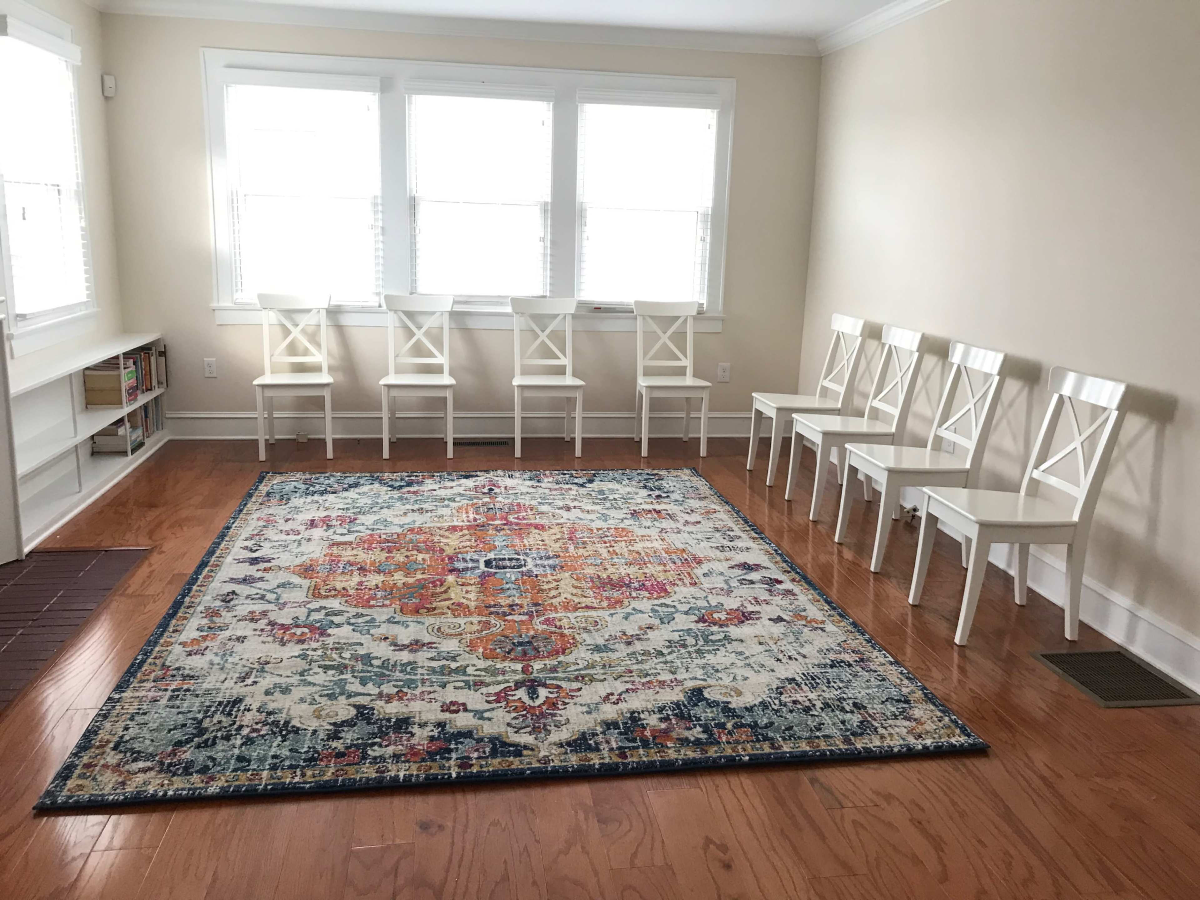 A sunlit room with a large, colorful area rug in the center and six white chairs positioned around it.