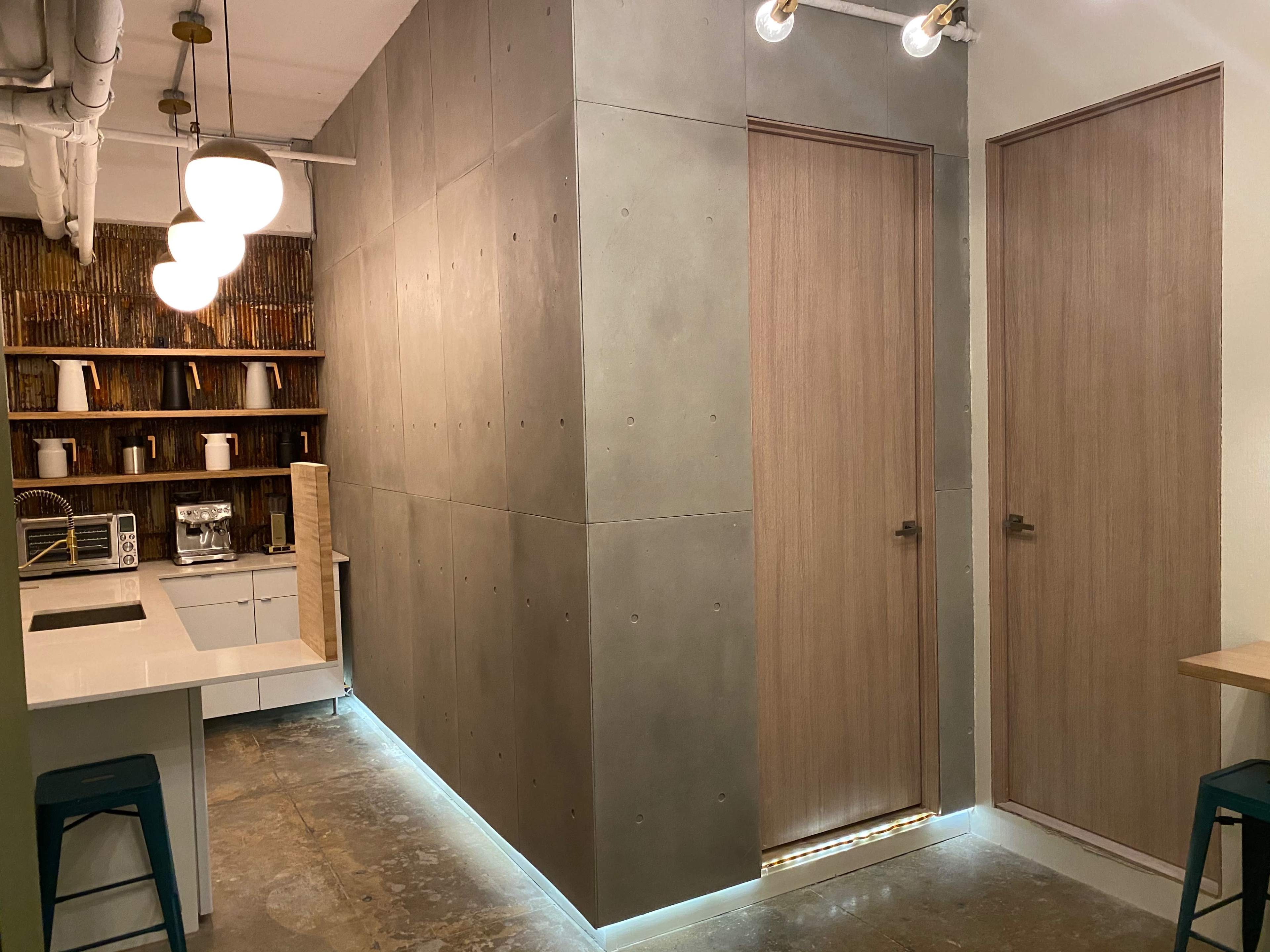 A modern interior space featuring a concrete wall with two wooden doors, a small kitchen area with a coffee machine, and shelves lined with white cups.