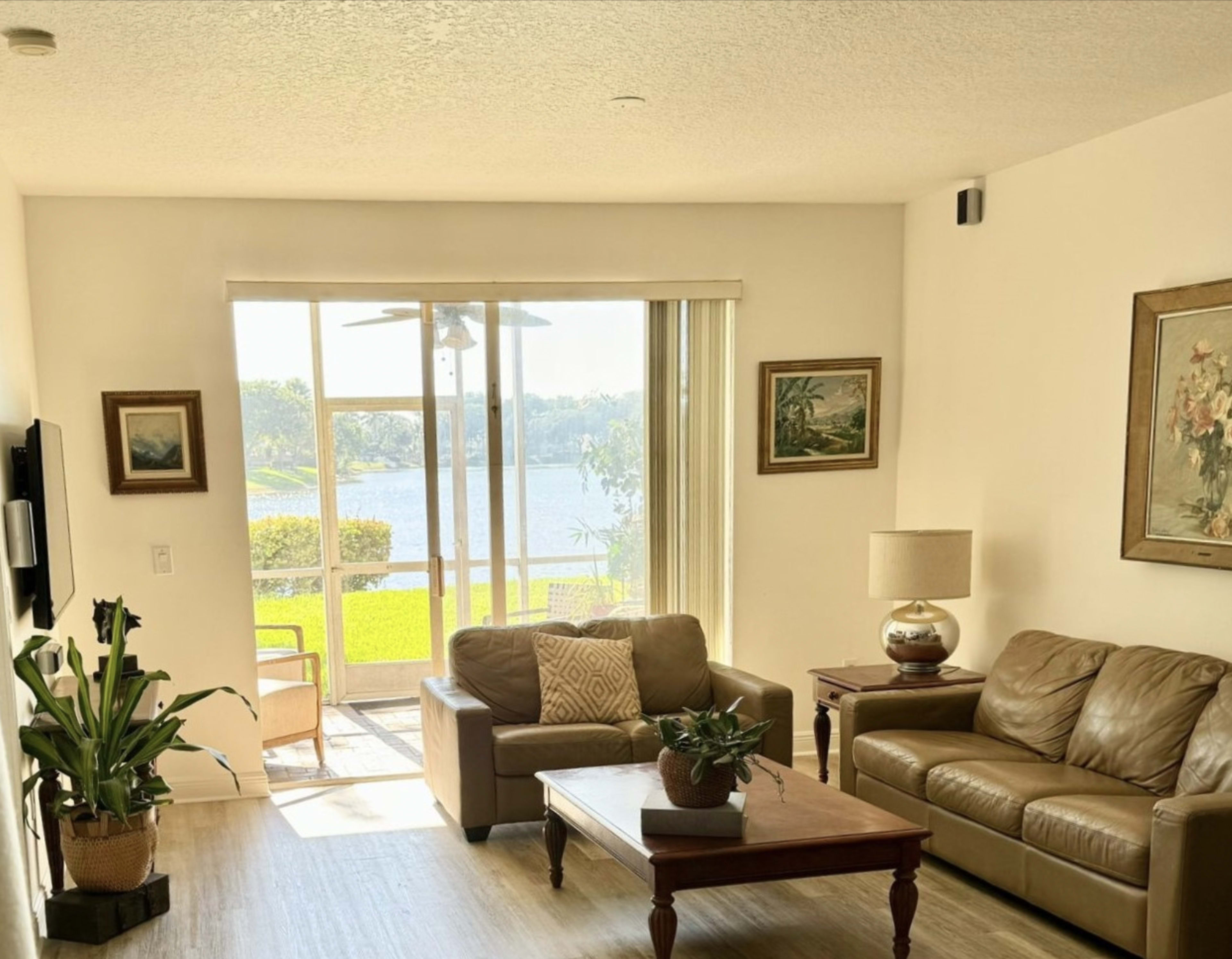 The image shows a bright living room with two brown couches, a wooden coffee table, and a view of a lake through large windows.