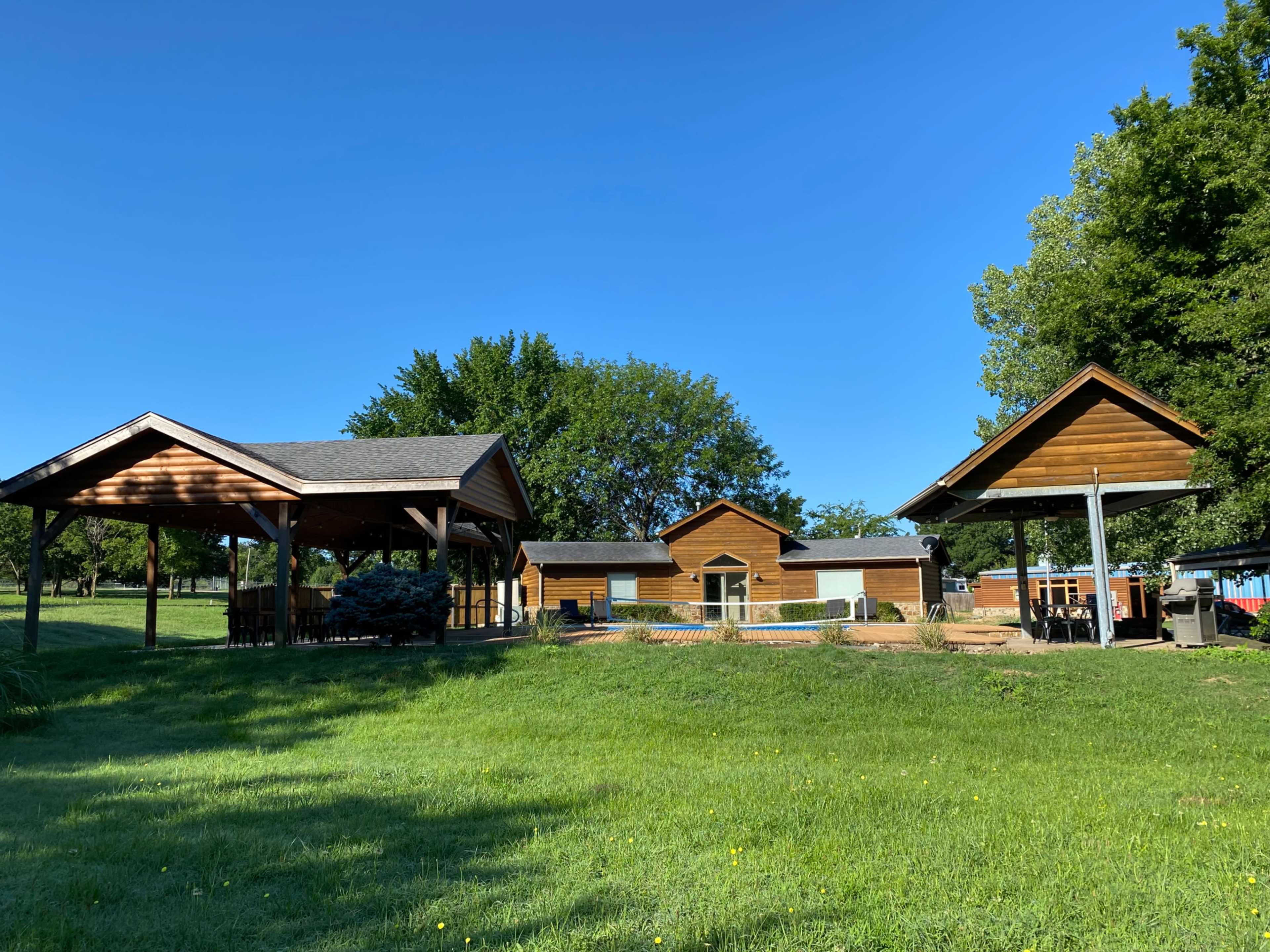 A wooden building complex with two covered structures on a grassy area under a clear blue sky.