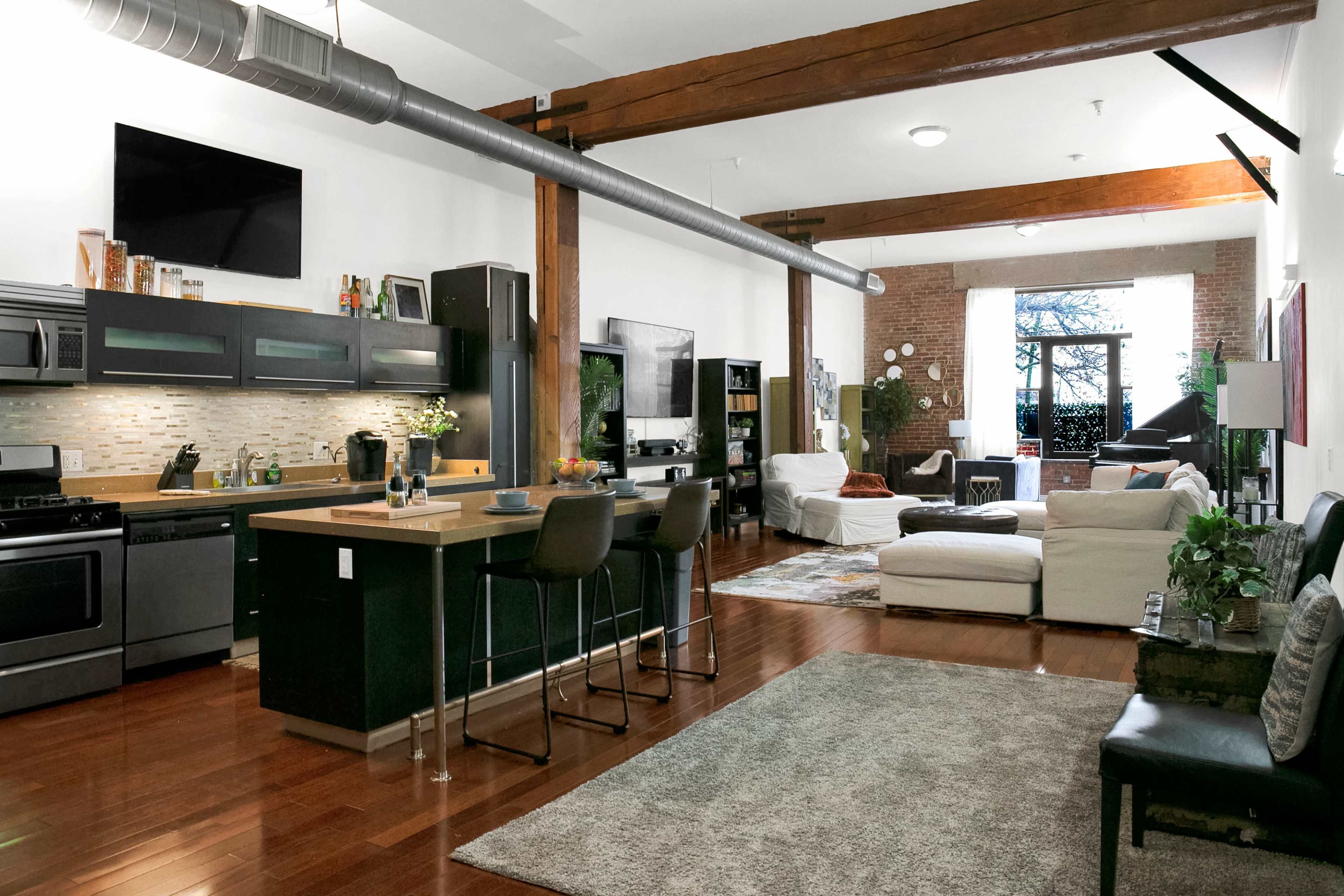 The image depicts a spacious, open-concept living area featuring a kitchen with a large island, modern appliances, a seating area with a sofa and armchair, and large windows allowing natural light to fill the room.