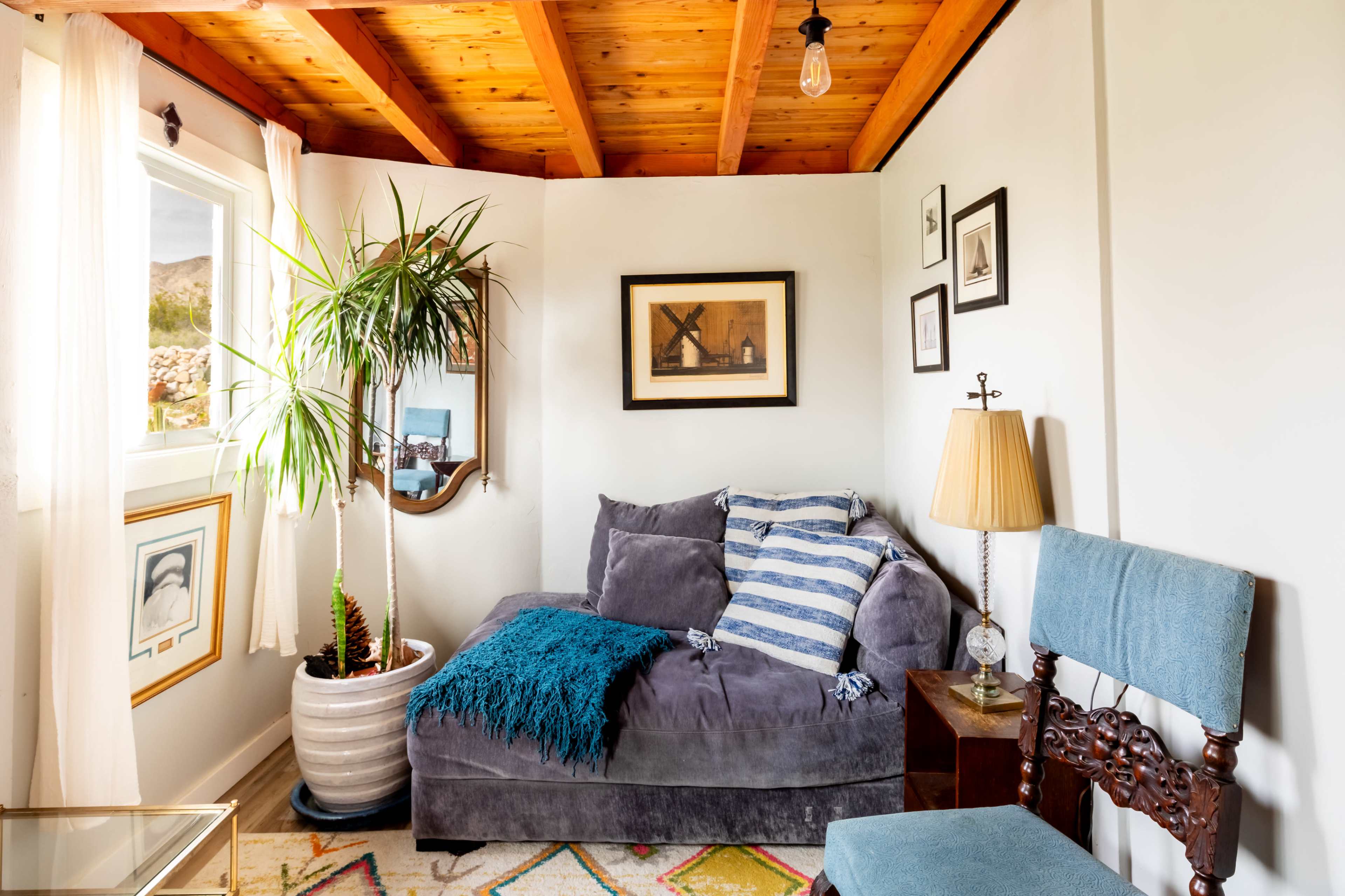 A cozy corner of a room features a gray couch adorned with blue and white striped pillows, a potted plant, and framed pictures on the walls, complemented by a wooden ceiling and a nearby lamp.