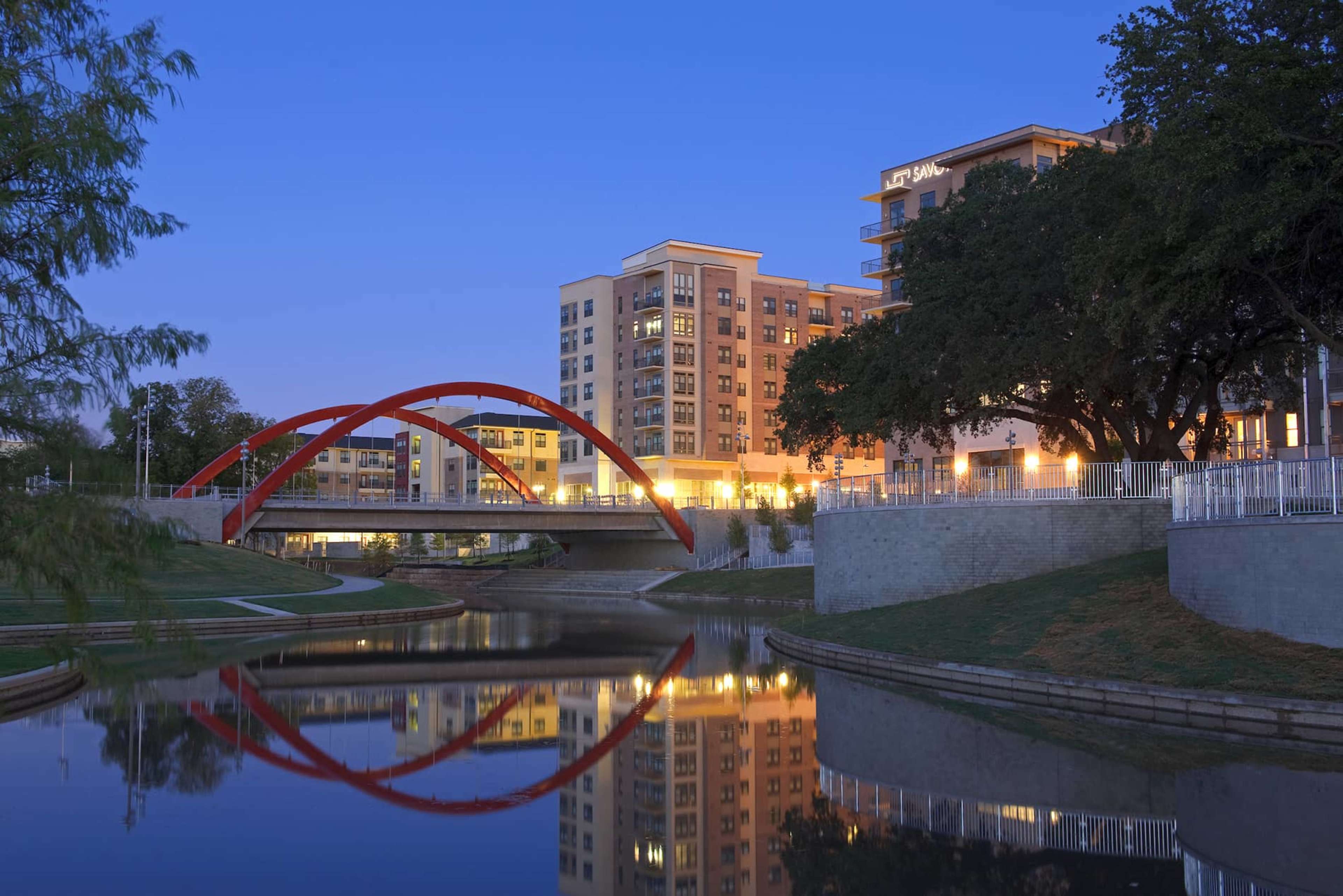 A modern apartment building with large windows stands beside a serene waterway, featuring a red arch bridge reflected in the water at dusk.