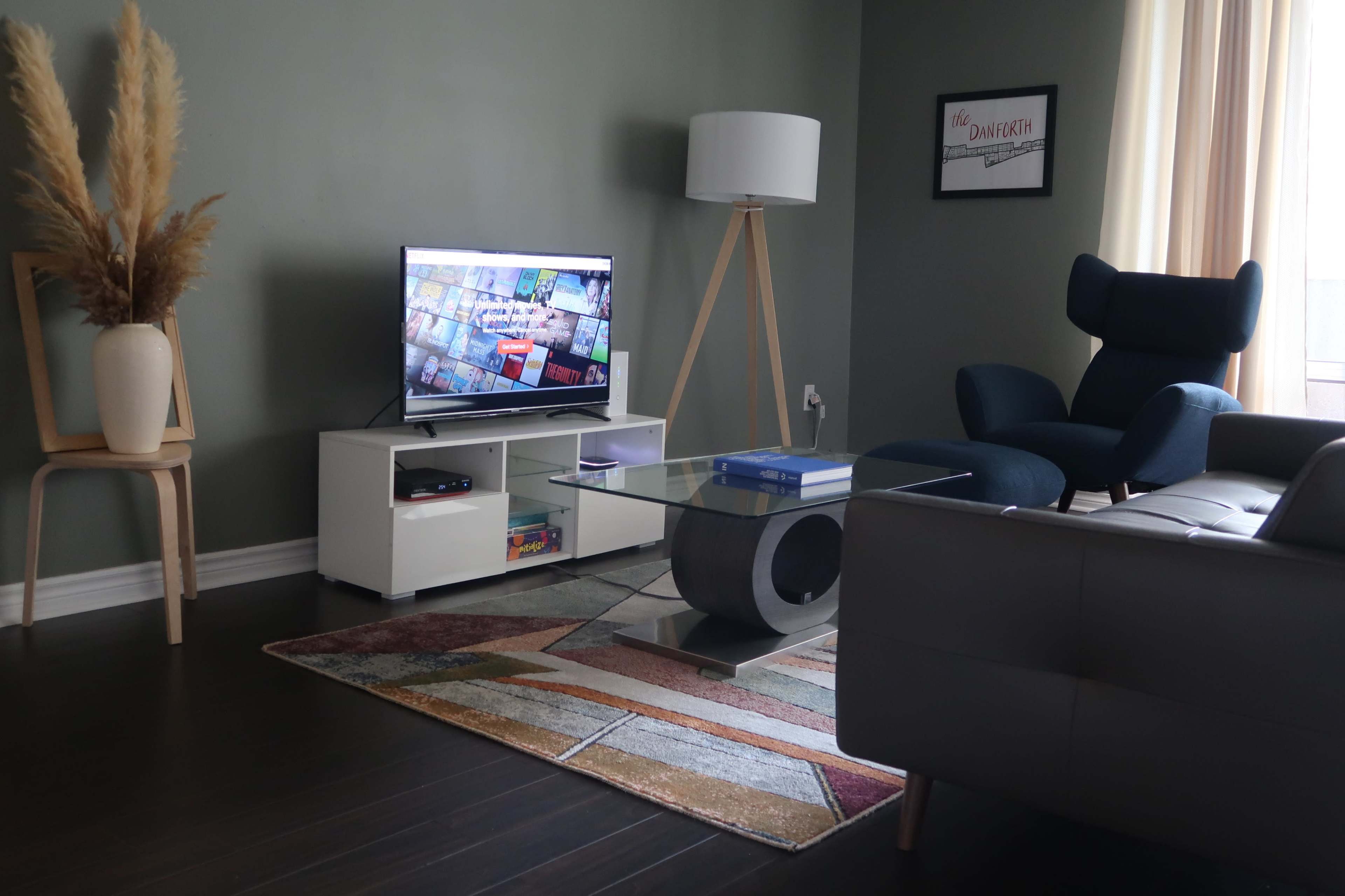 A modern living room features a television on a white stand, a blue chair, a glass coffee table, and a decorative vase with pampas grass against a green wall.