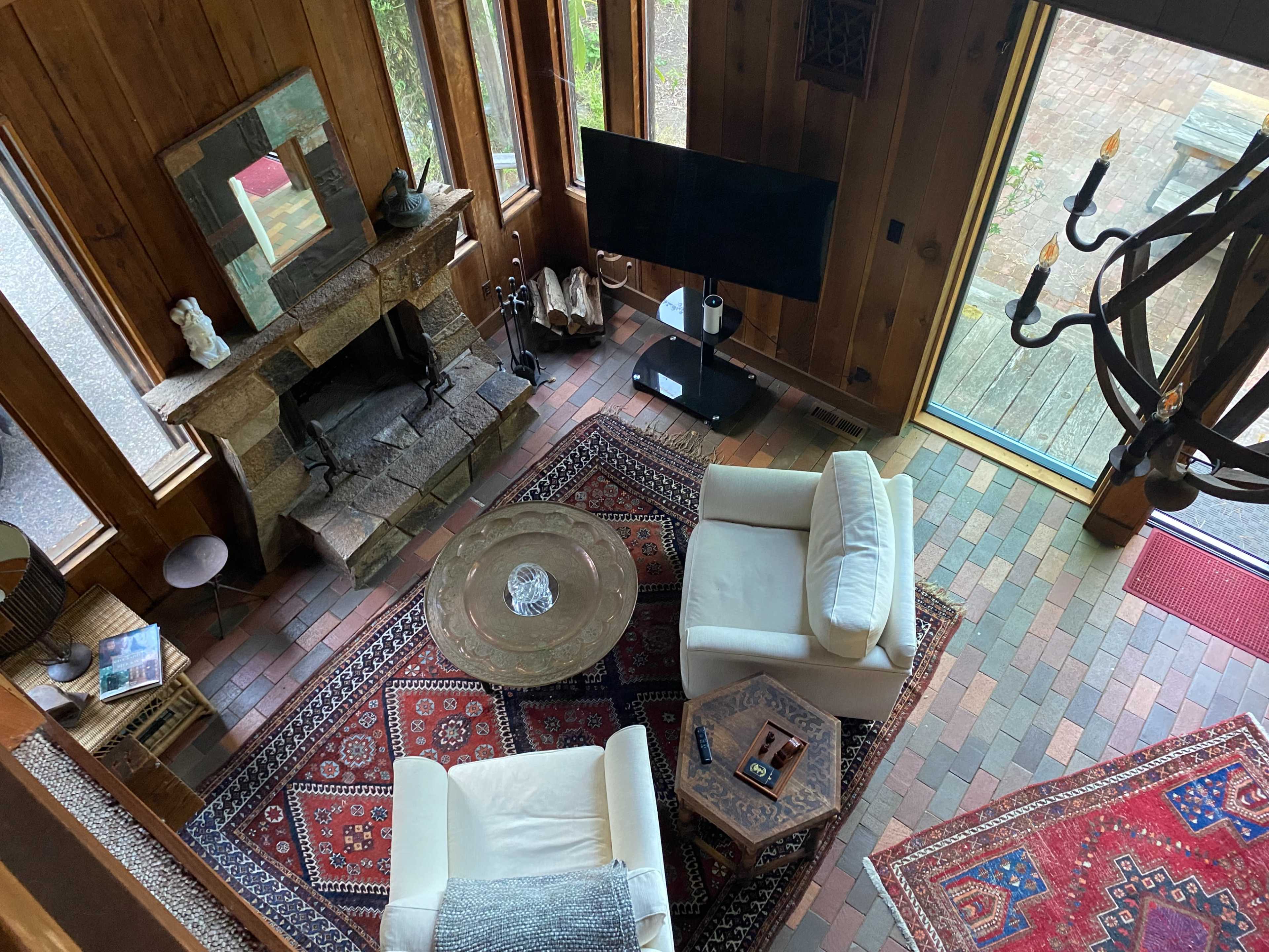 A wooden living room with two white chairs, a round coffee table, a stone fireplace, and a large television set against a wall of windows.