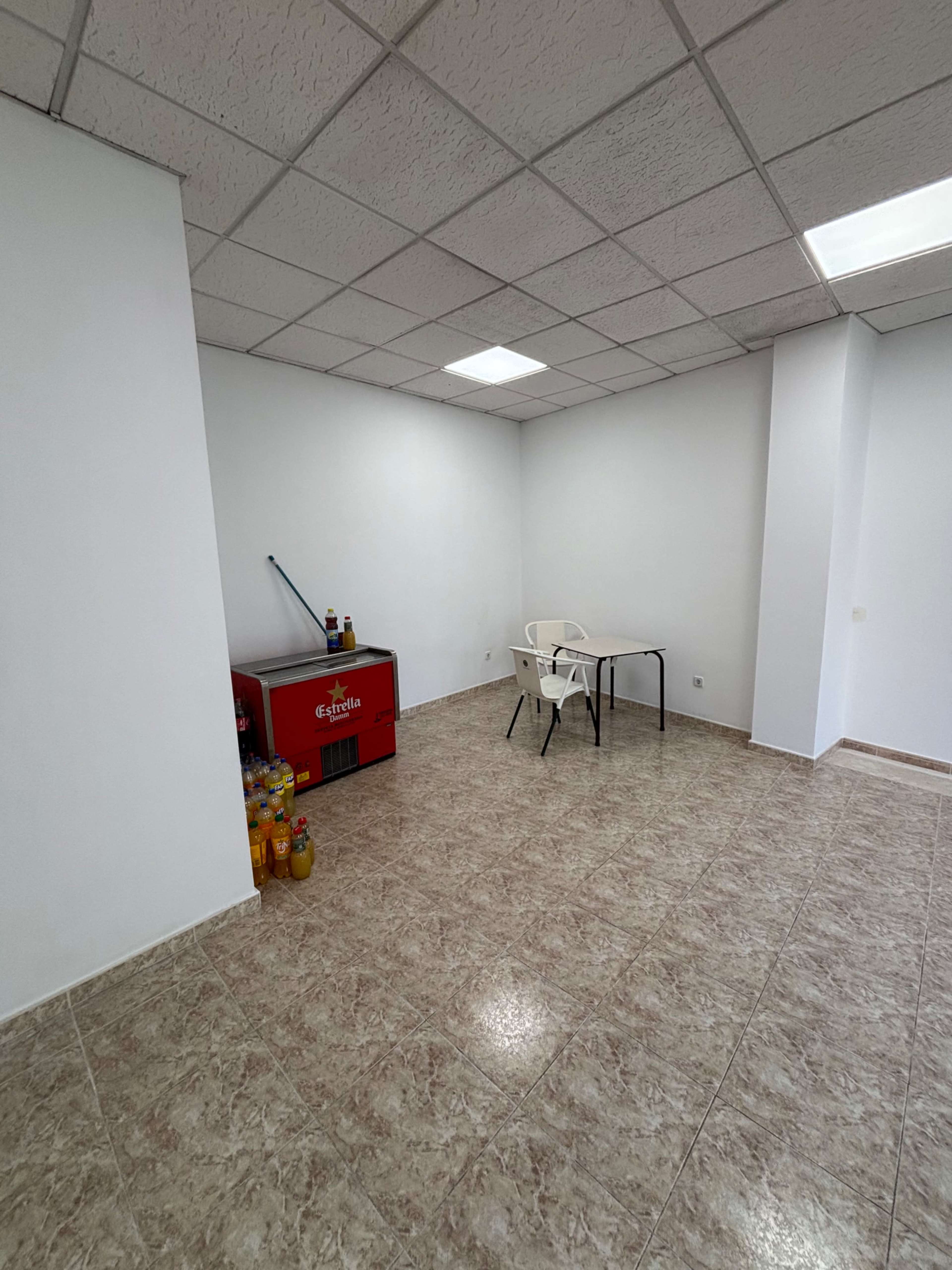 The image shows a minimally furnished room featuring a table with two chairs, a red cooler beside it, and shelves with various drink bottles.