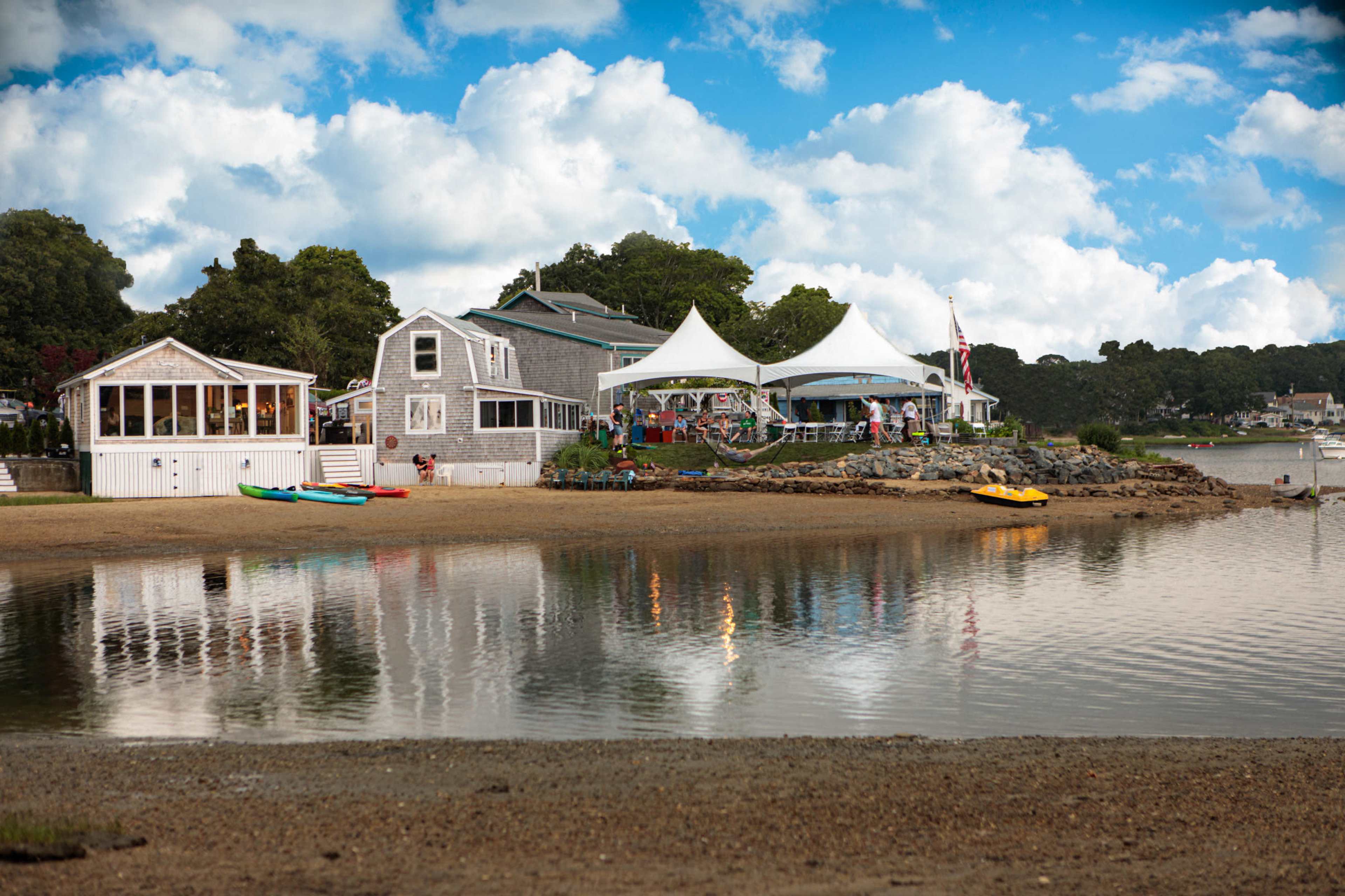 A calm waterfront scene with several houses, a large white tent, and colorful kayaks on the shore.