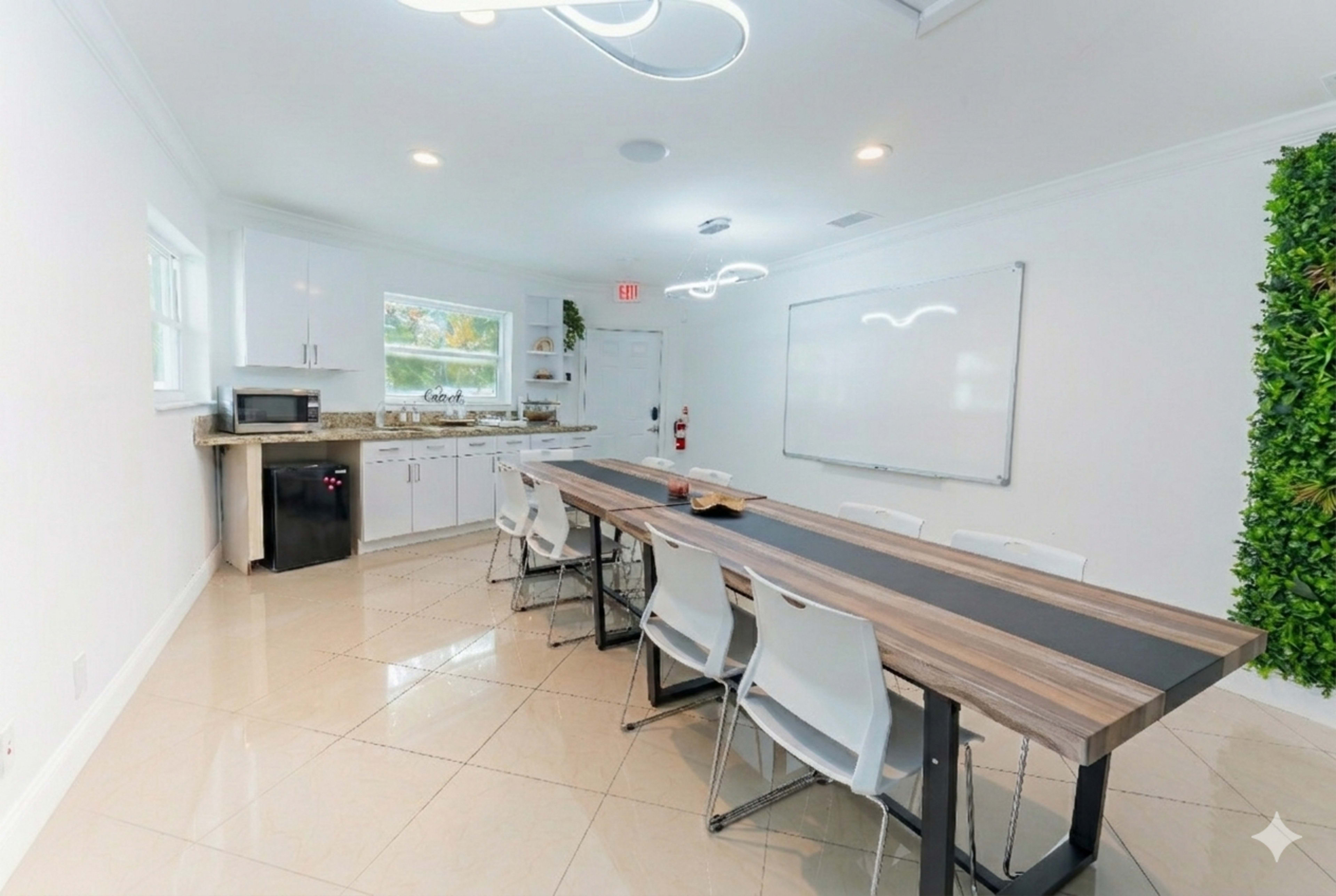 The image shows a modern meeting room with a long wooden table, white chairs, a kitchenette, and a whiteboard on the wall.