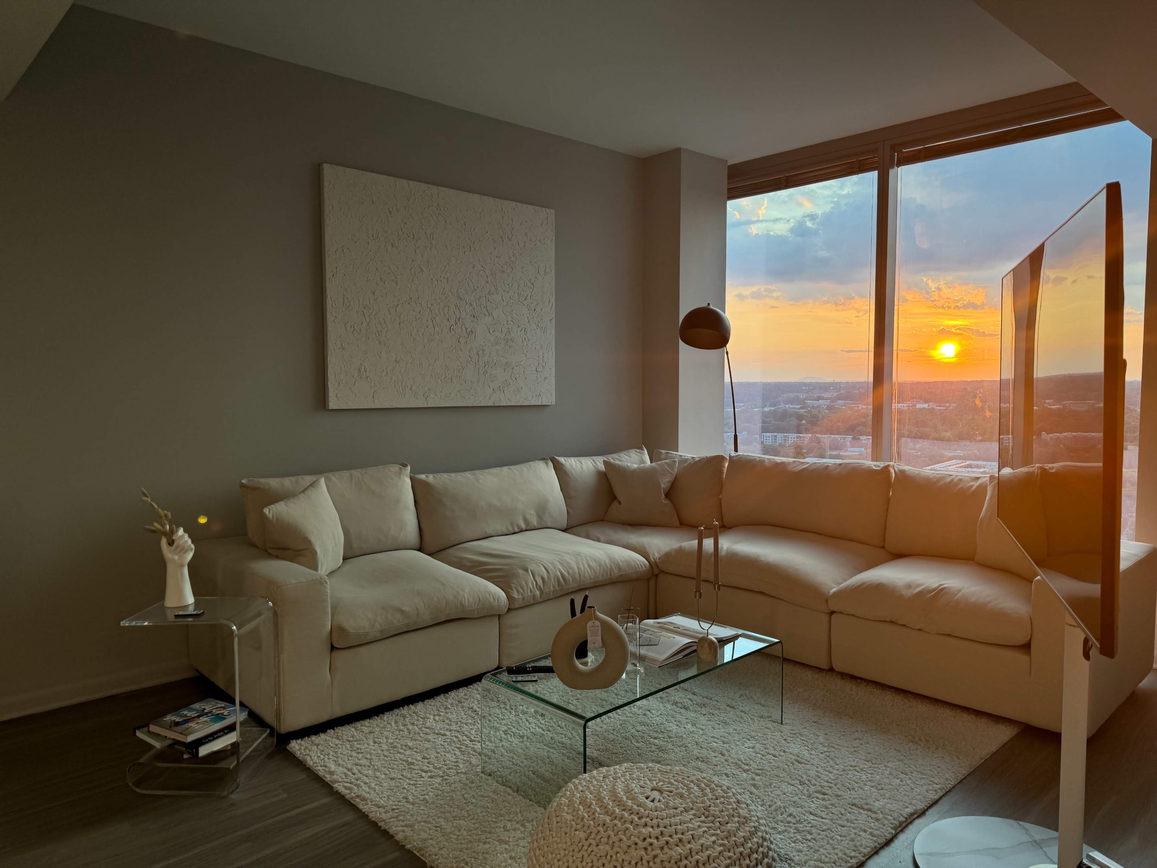 A spacious living room features a large beige sectional sofa, a round coffee table, and a floor lamp, with a view of a sunset visible through a large window.