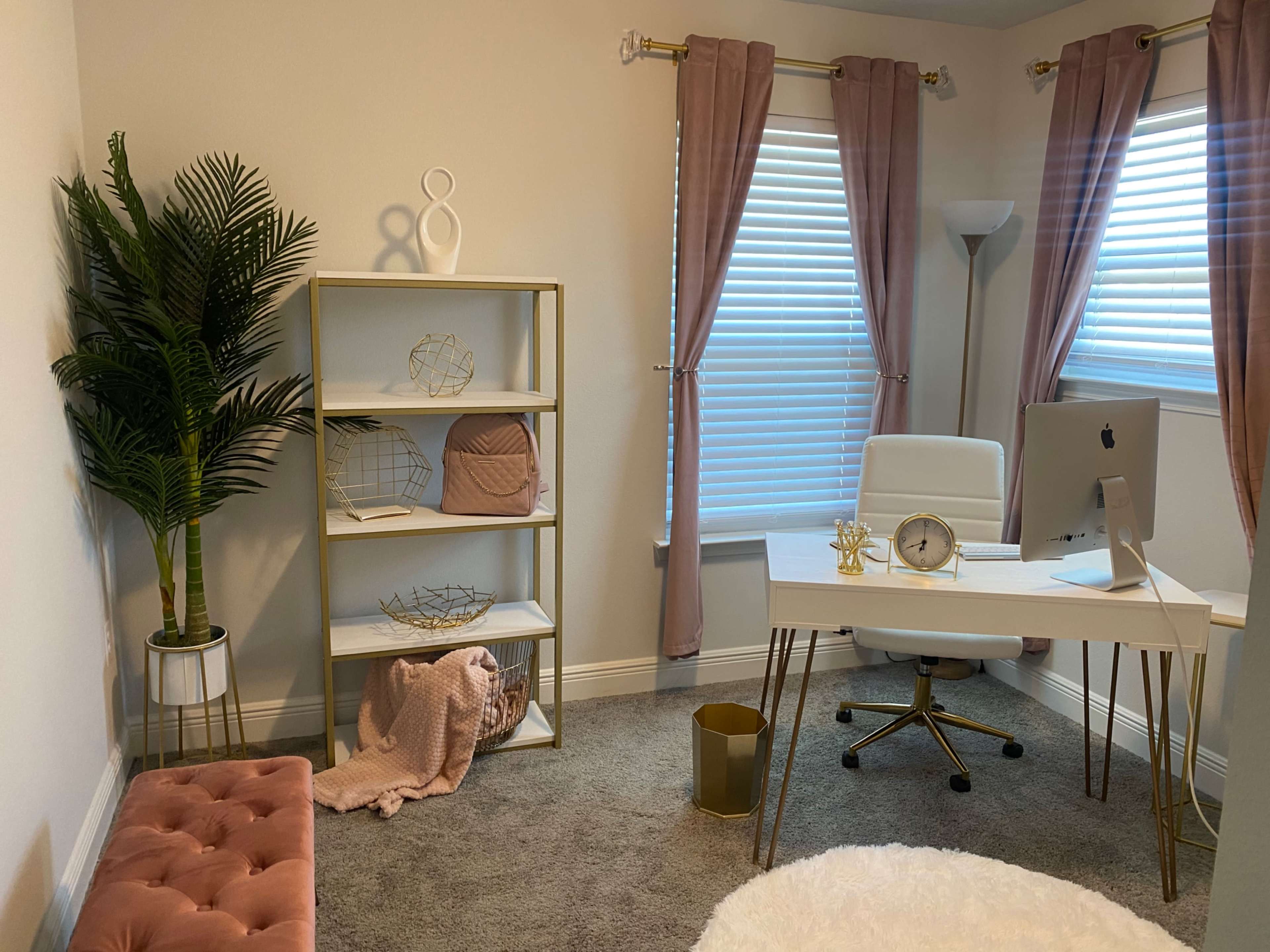 A modern home office with a white desk and chair, a tall bookshelf filled with decor, and large windows covered with pink curtains.