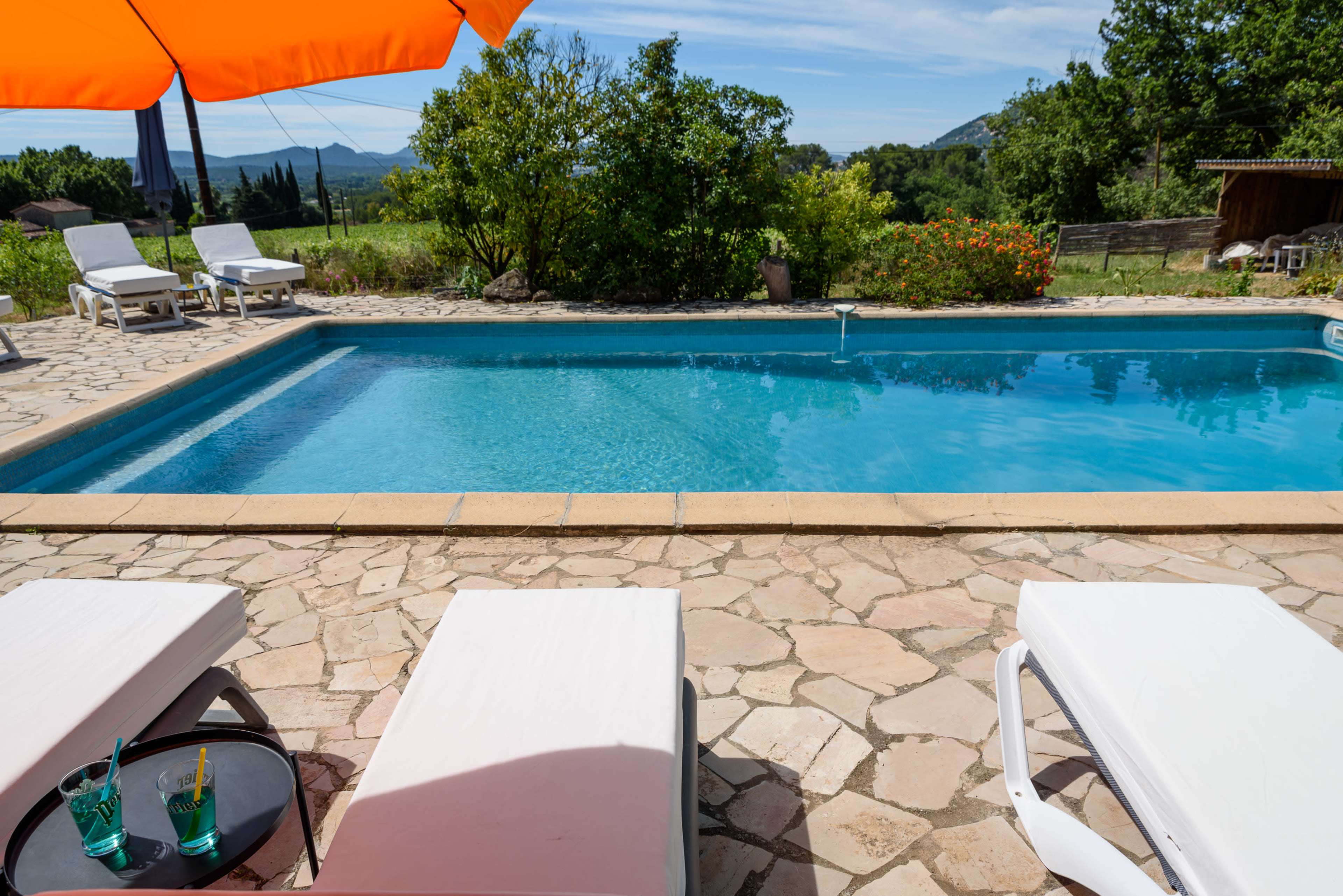 A crystal-clear swimming pool is surrounded by lounge chairs and vibrant greenery under a bright orange umbrella.