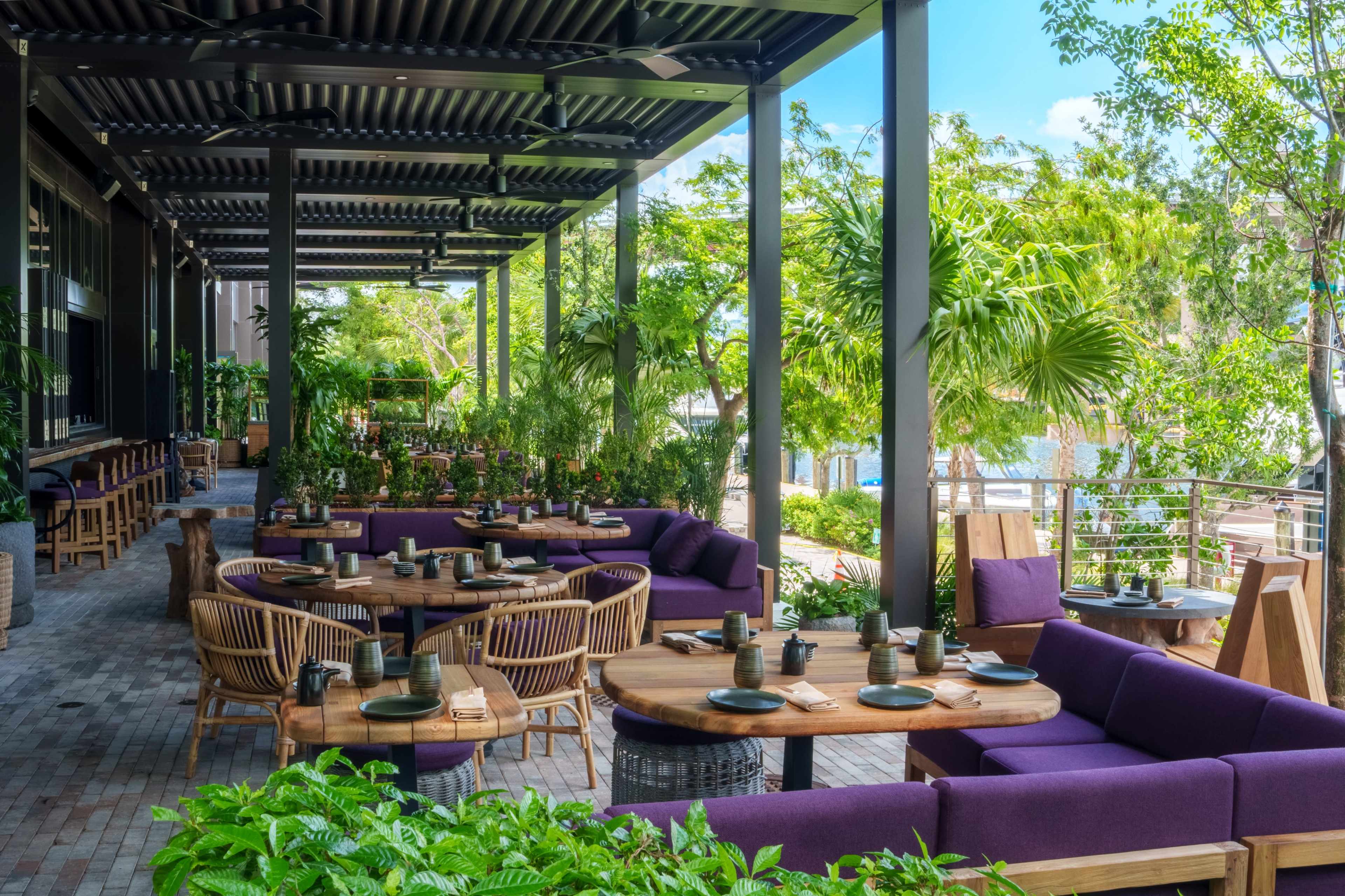A modern restaurant patio features wooden tables surrounded by purple seating and lush greenery.