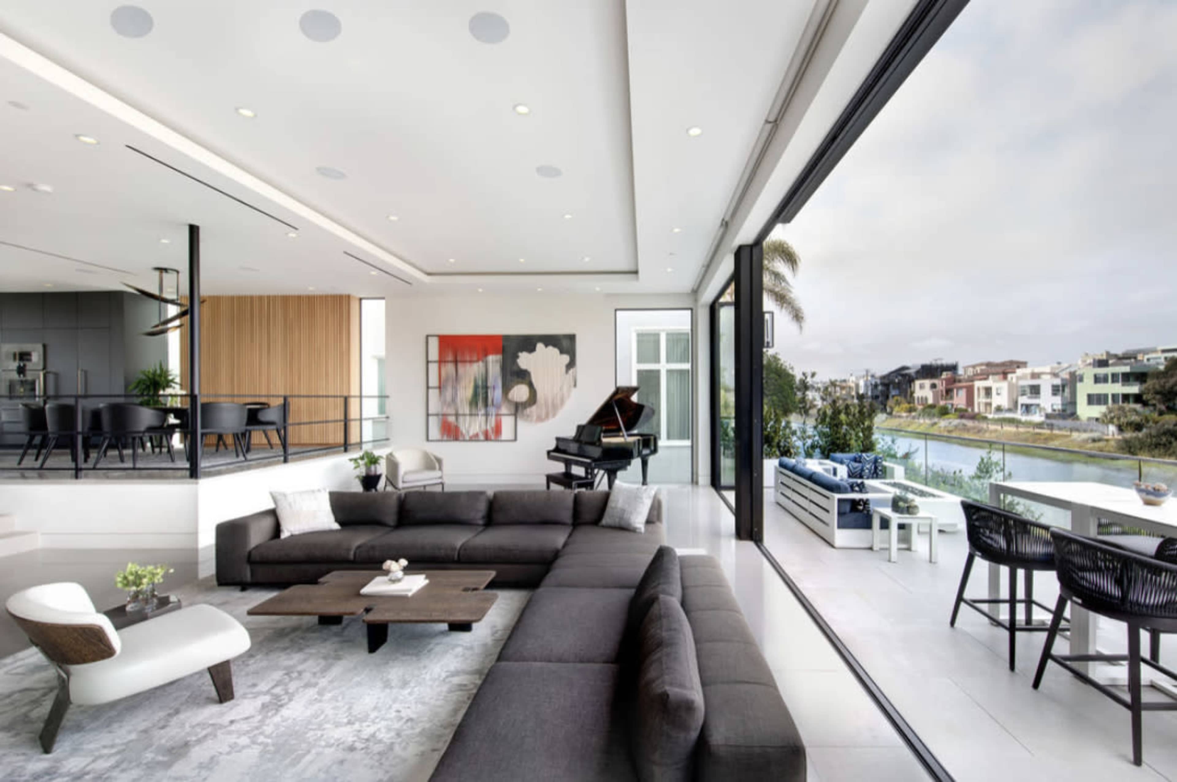 A modern, open-concept living space featuring a large gray sectional sofa, a piano, and views of a waterfront through floor-to-ceiling windows.