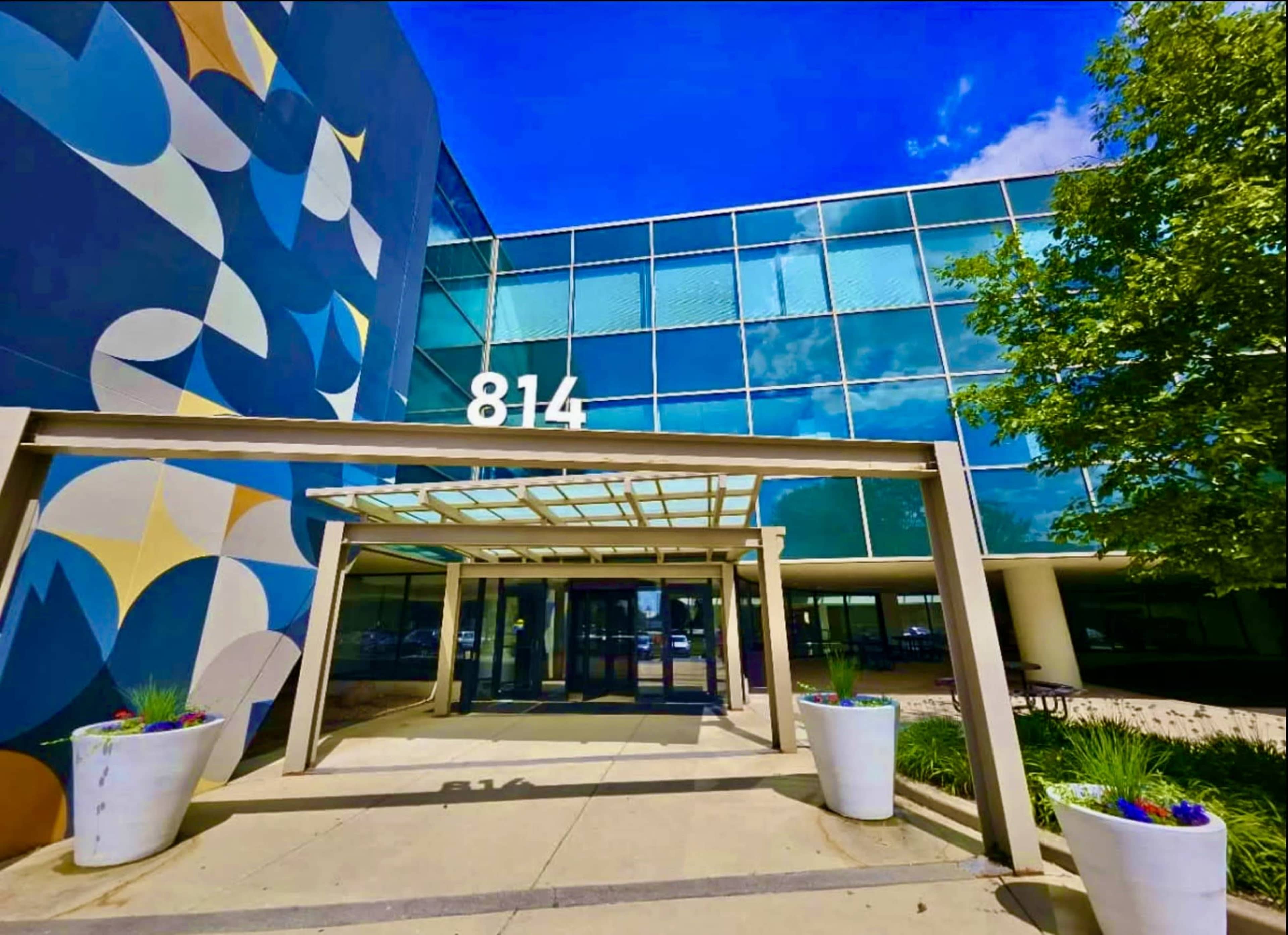 The image shows a modern building entrance with a large blue and abstract patterned wall, a glass facade, and a canopy over the walkway.
