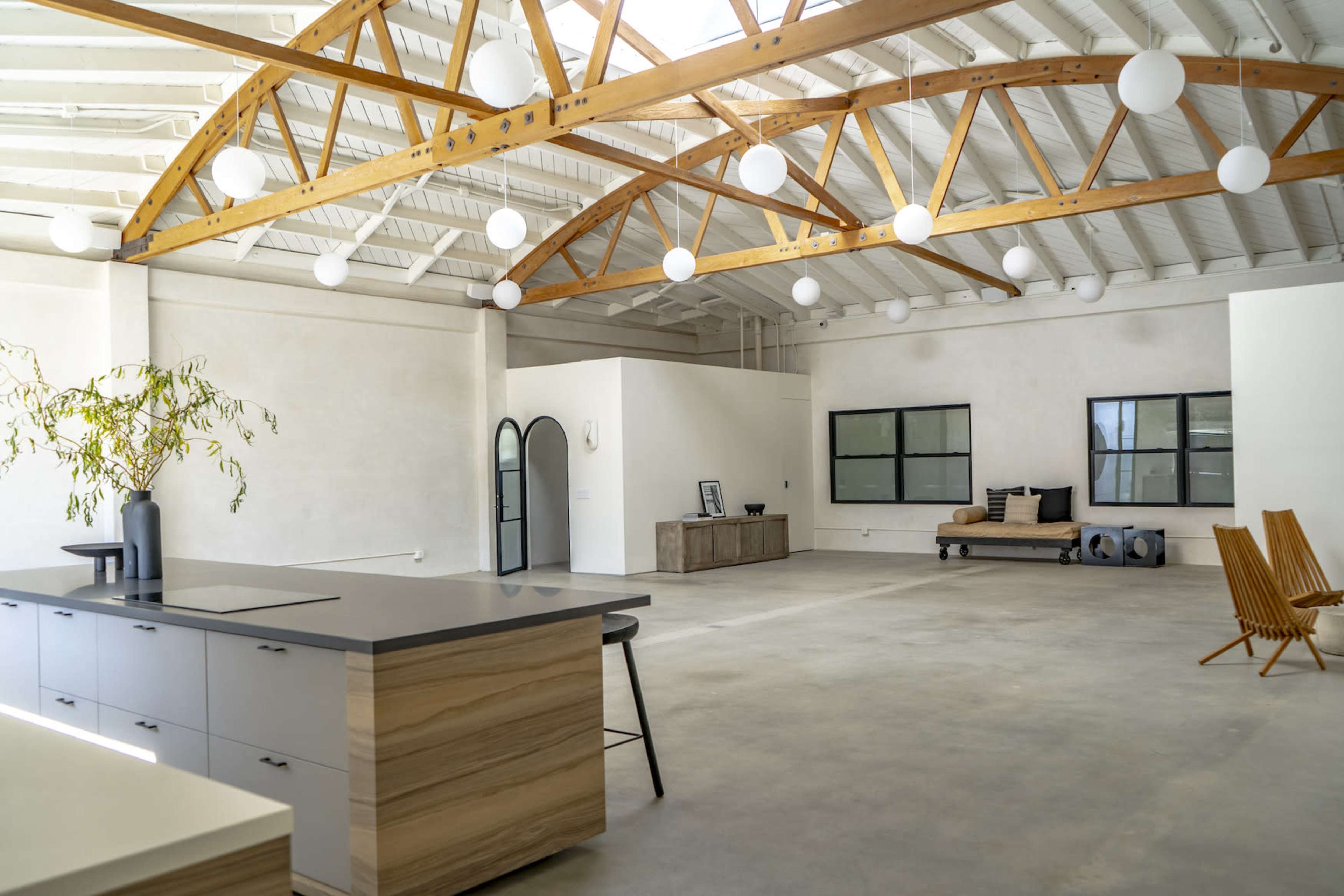 A spacious room with high ceilings, wooden beams, and a minimalist kitchen island, featuring a lounge area with a sofa and decorative plants.