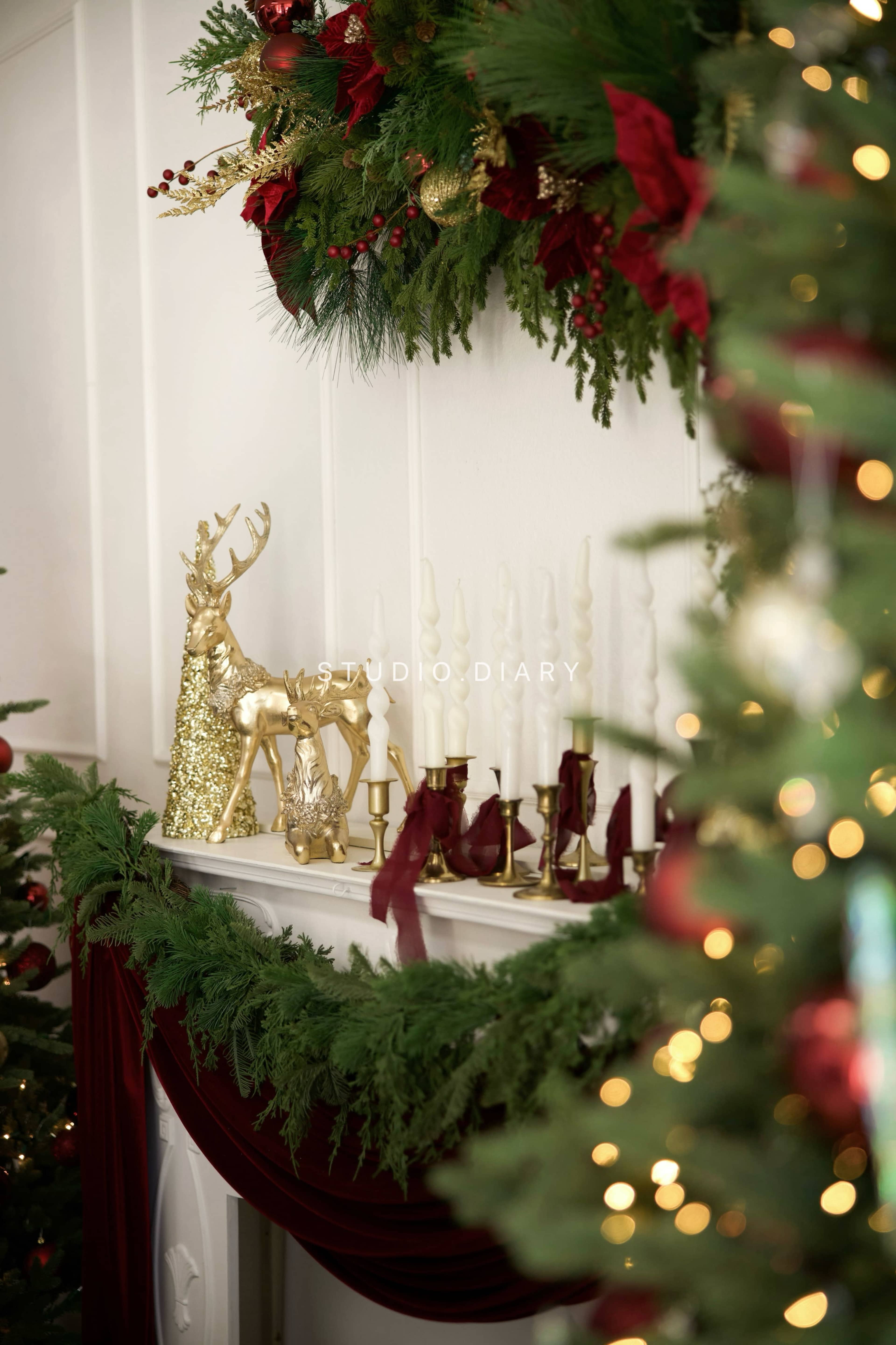 The image depicts a festive holiday mantel adorned with greenery, gold decorative deer, and candles, all complemented by rich red accents.