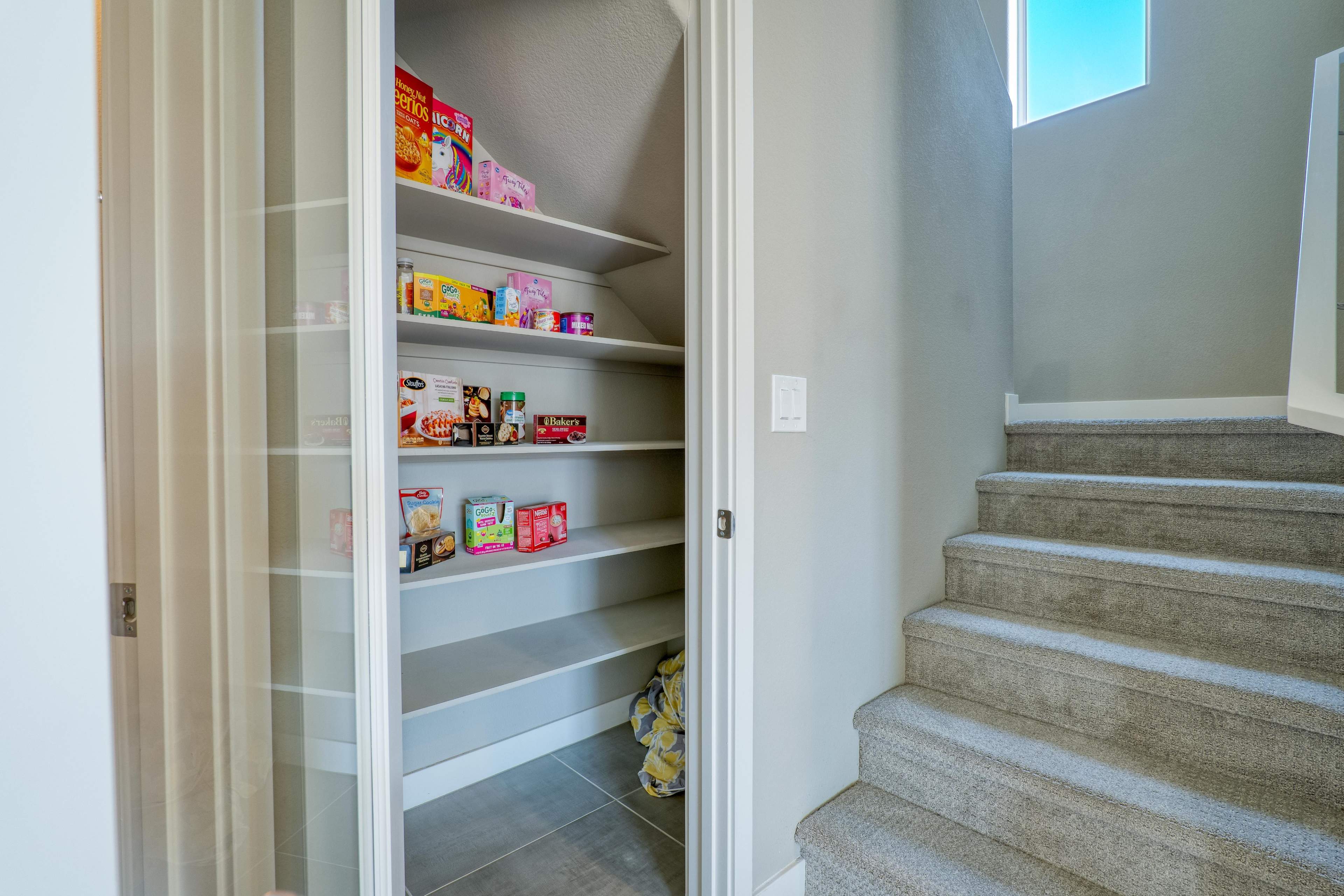 A pantry with several shelves containing various food items is located beside a staircase.