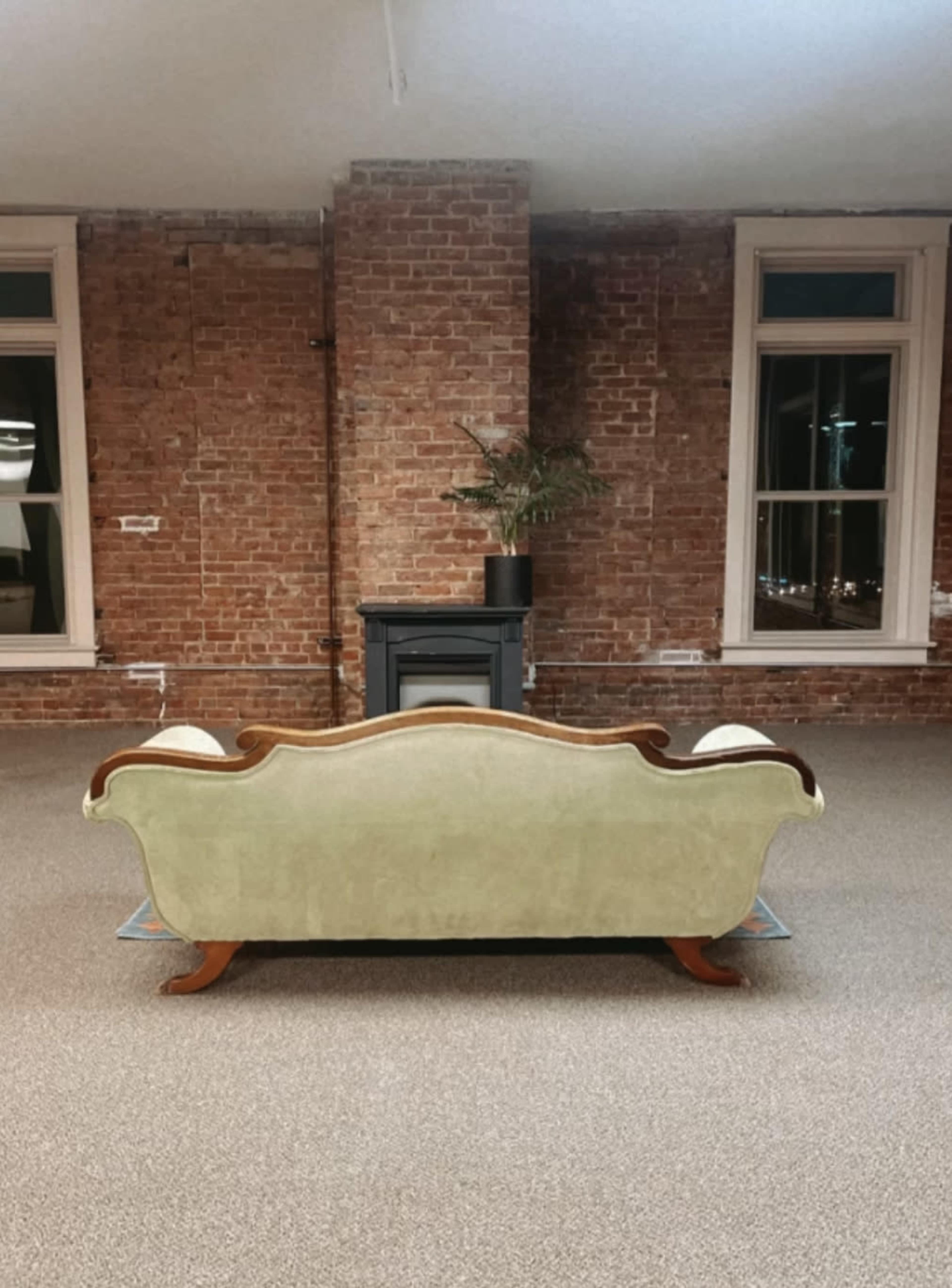 A vintage green sofa is positioned against a red-brick wall in a spacious room with large windows.