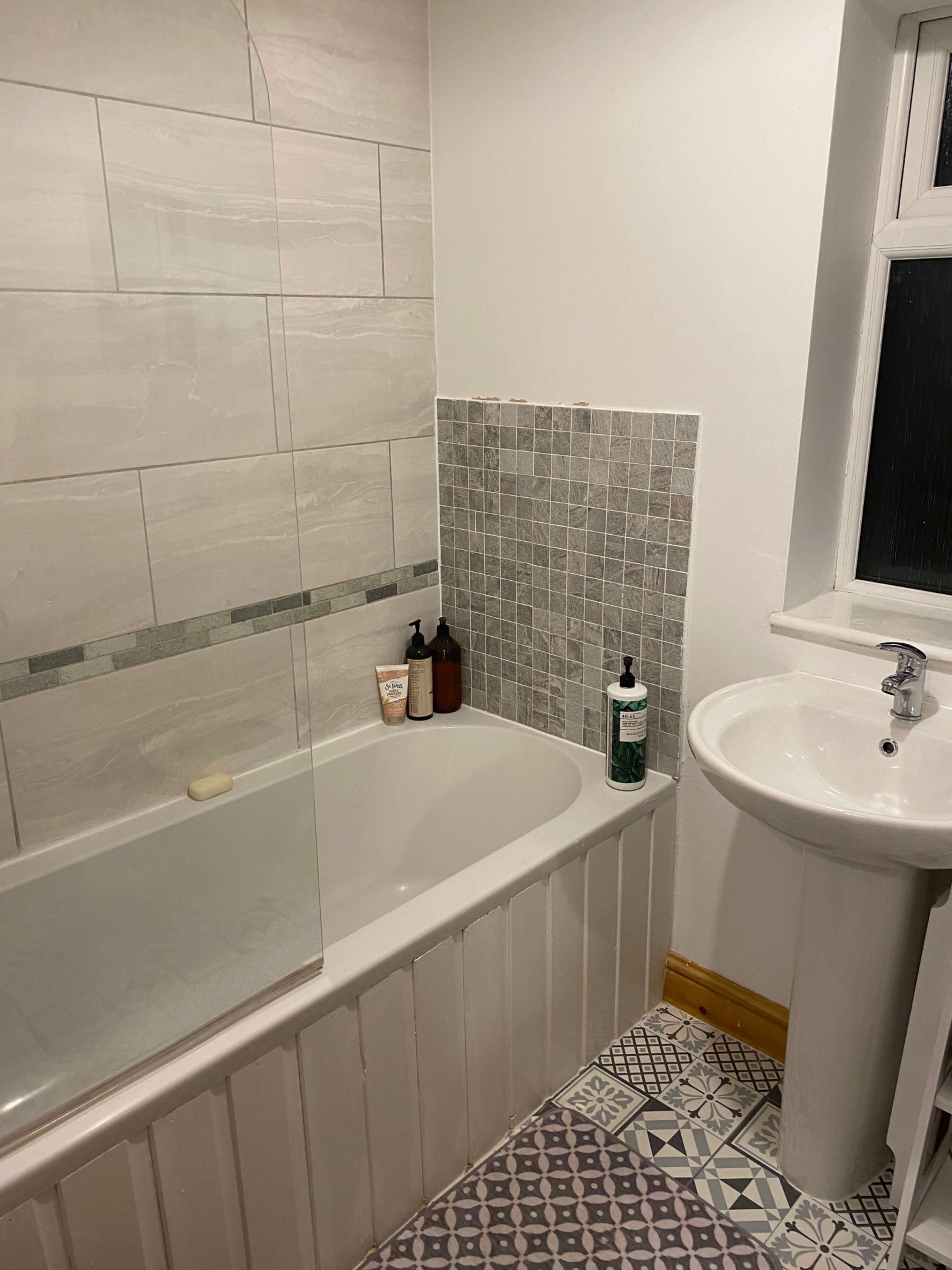 The image shows a bathroom featuring a bathtub with a glass screen, a pedestal sink, and patterned tiles on the floor.