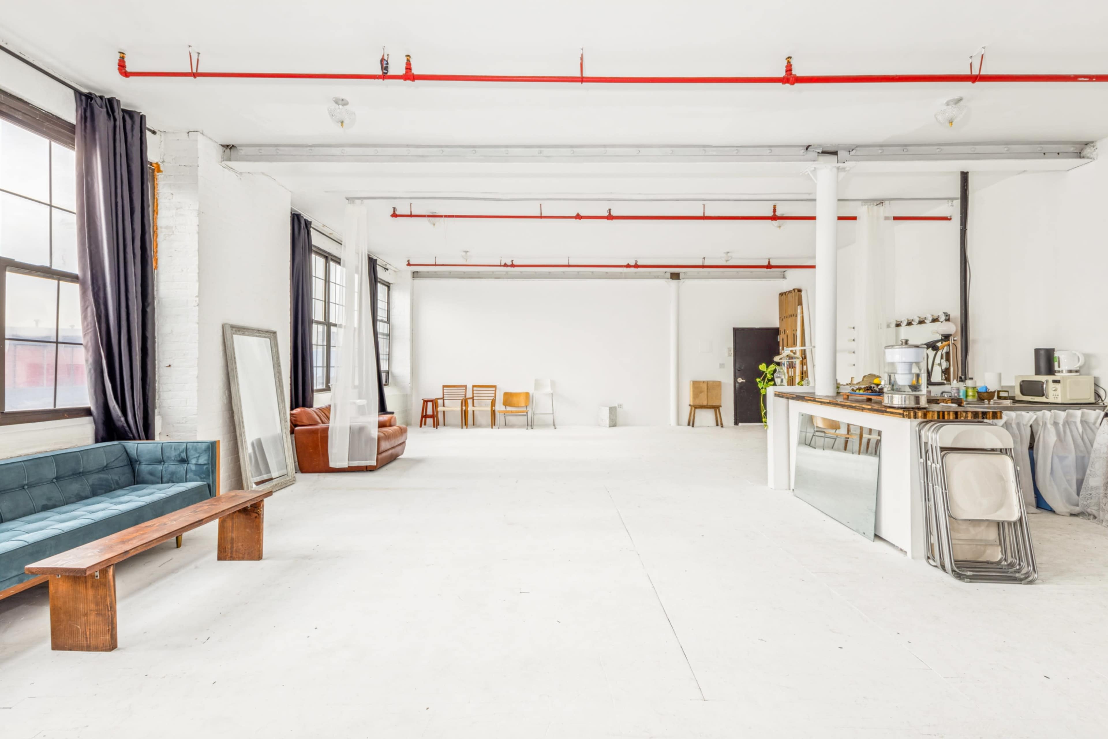 An expansive, minimalist studio space features large windows, a wooden bench, a brown sofa, various chairs, and a kitchenette against a white backdrop.