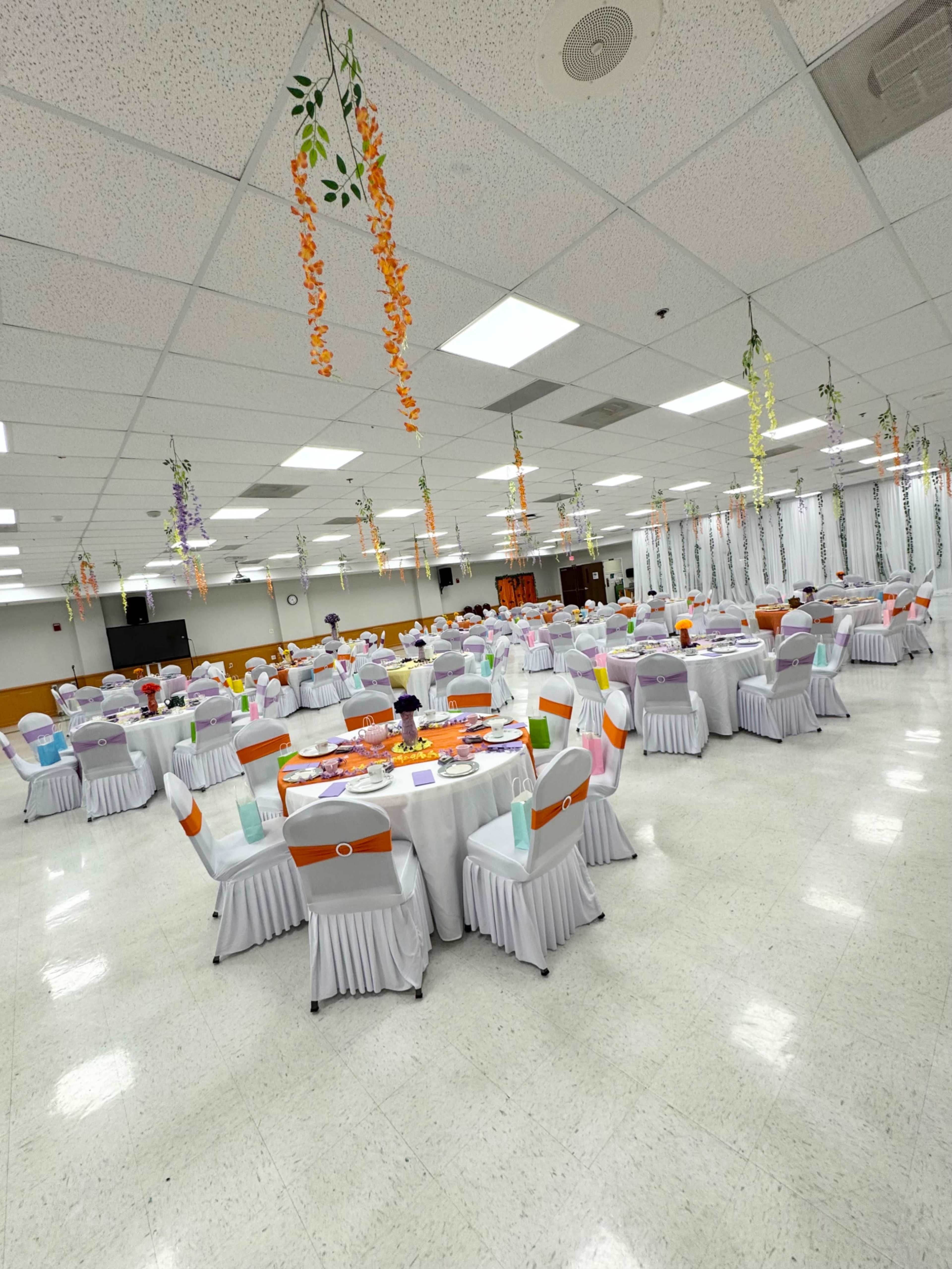 The image shows a large banquet hall set up with round tables covered in white tablecloths, decorated with colorful chair sashes and floral decorations hanging from the ceiling.