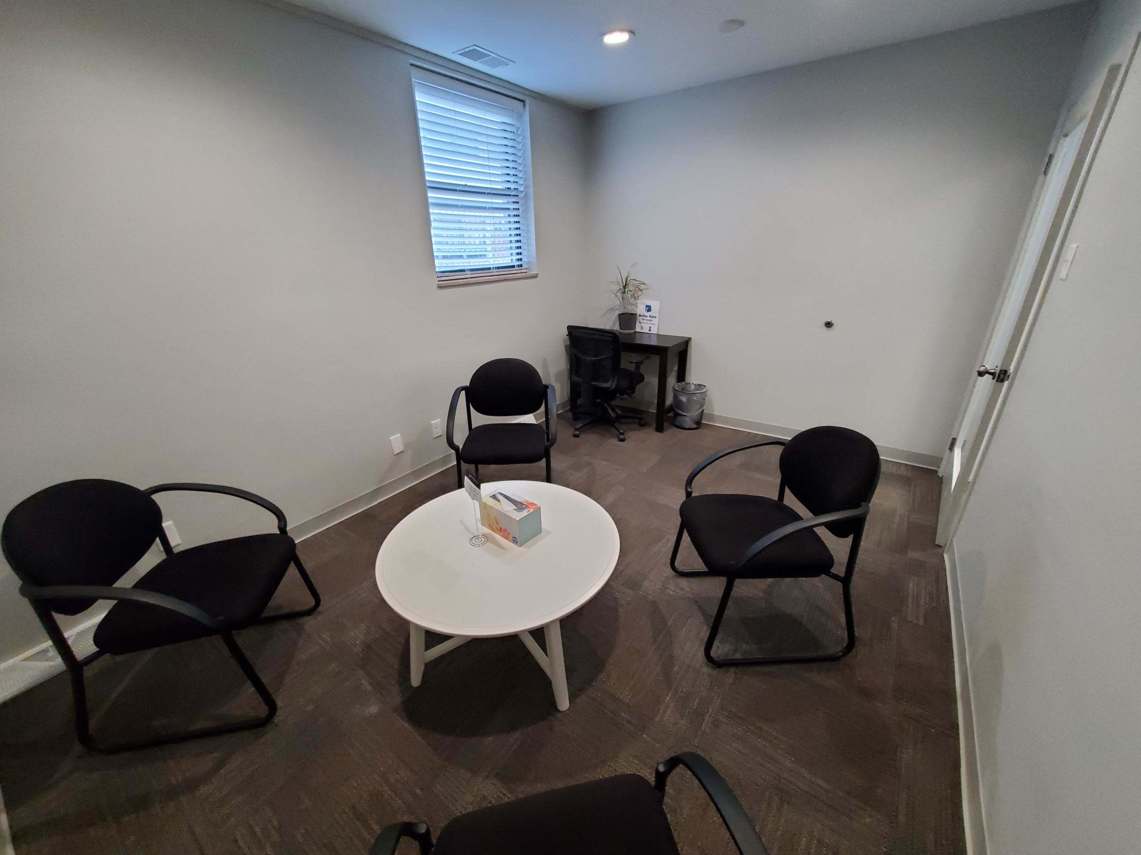 A small room features a circular table with a box on it and four black chairs arranged around it, along with a desk in the corner.