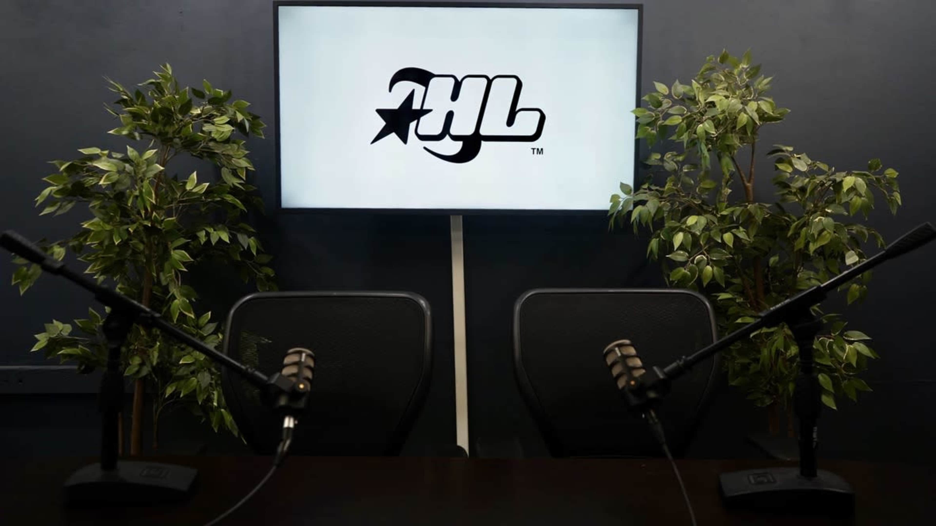 The image shows a small studio setup with two empty chairs facing a screen displaying a logo, surrounded by potted plants and equipped with microphones.