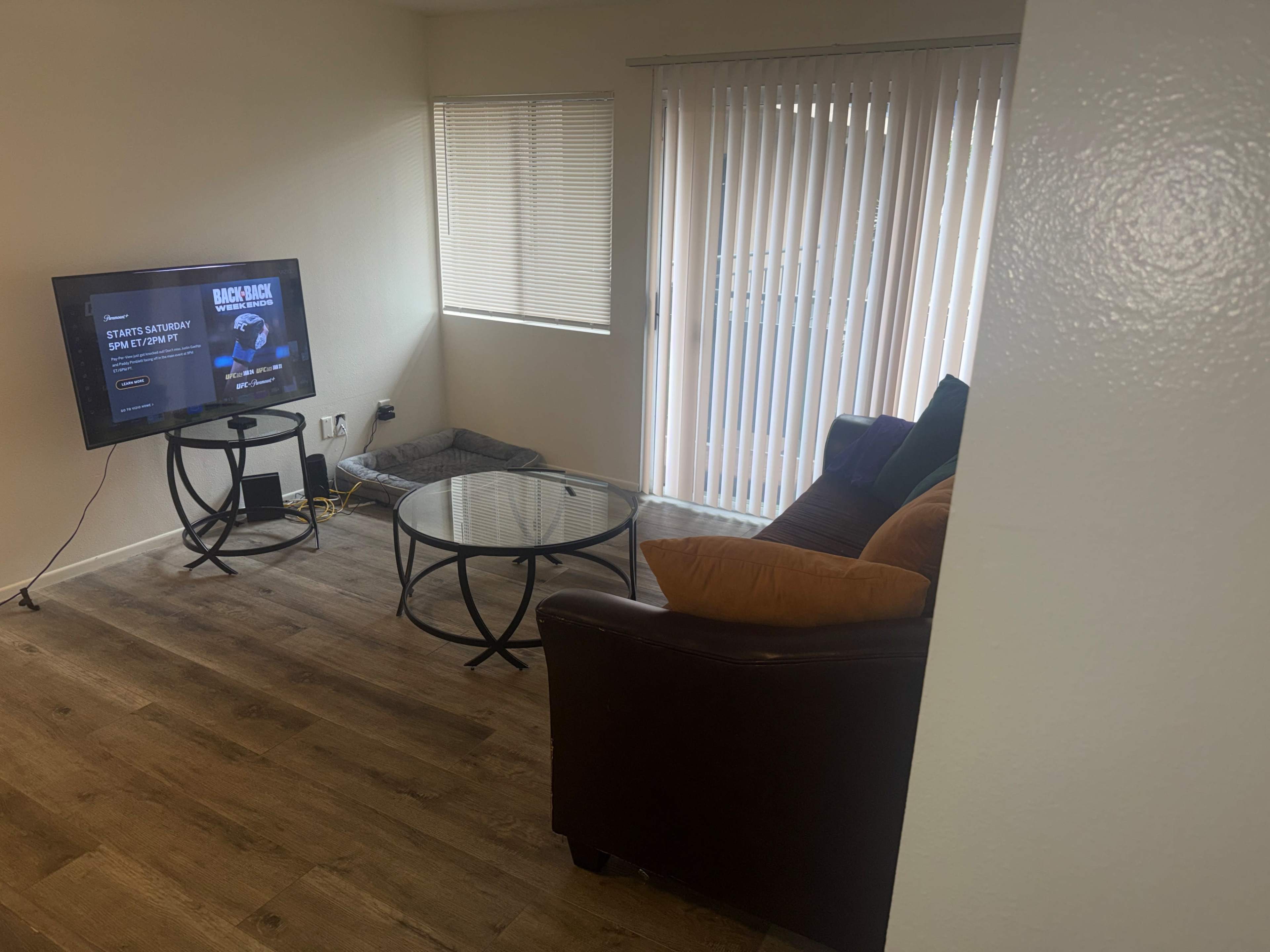 The image shows a living room with a television on a stand, a glass coffee table, a brown couch with orange cushions, and vertical blinds covering a window.