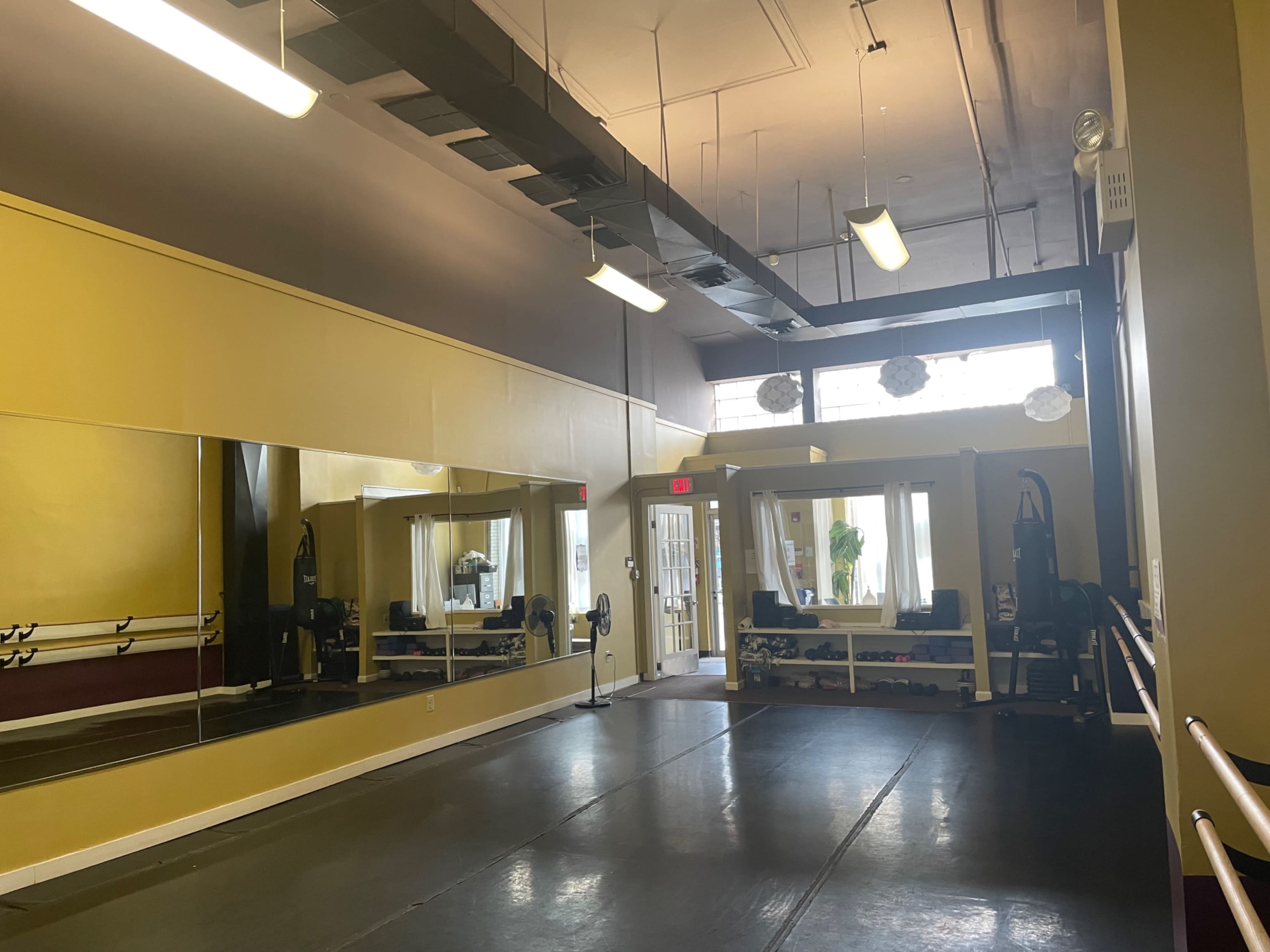 The image shows a spacious fitness studio with yellow walls, large mirrors, and exercise equipment arranged along one side.