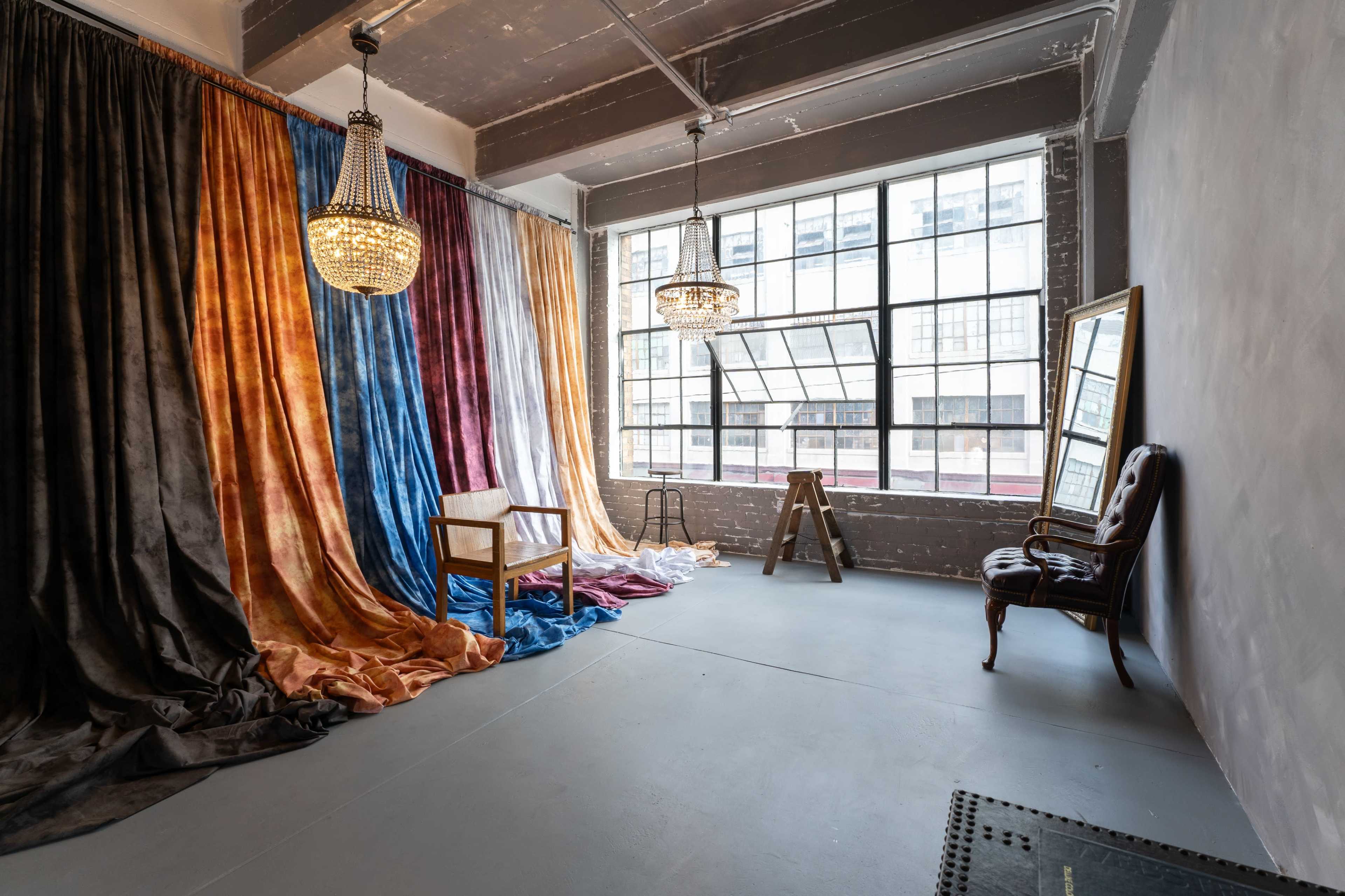 A spacious room with colorful fabric drapes, a mirror, a chair, and two chandeliers, illuminated by natural light from large windows.