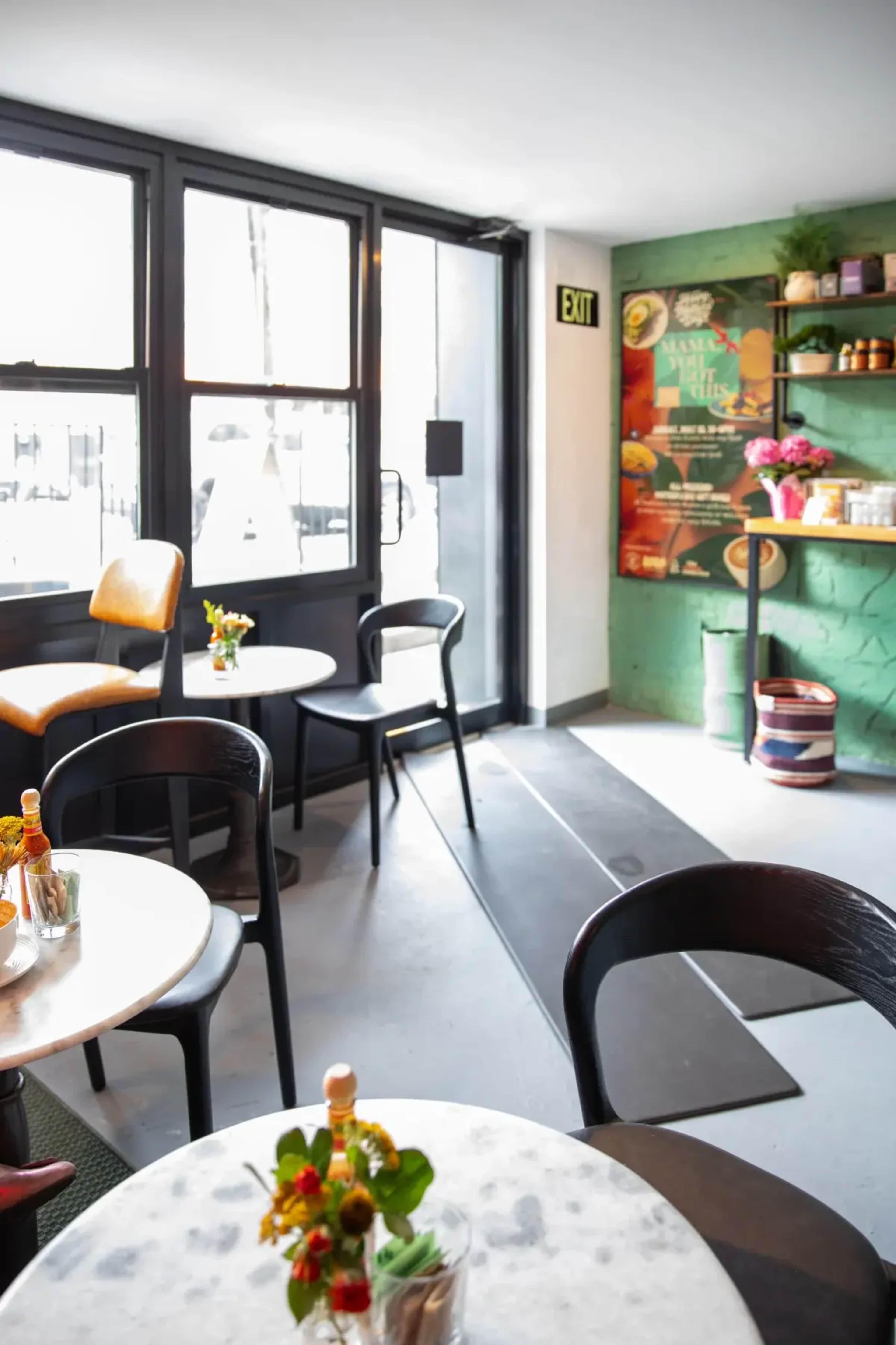 The image shows a cozy café interior with small round tables, black chairs, and a green accent wall featuring decorative items and menu highlights.
