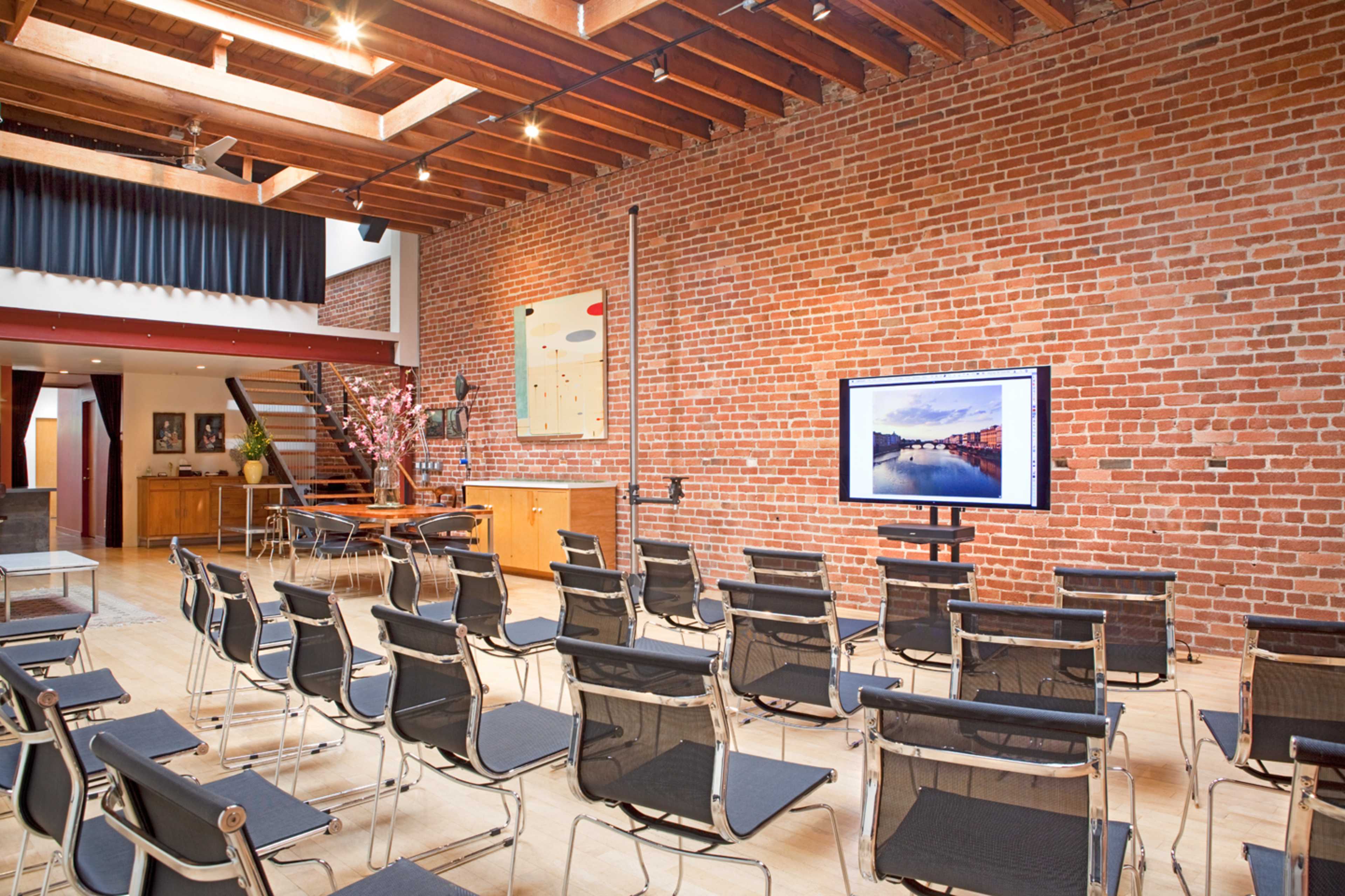 A modern conference room features rows of black chairs facing a large screen against a brick wall, with wooden beams overhead and an open space in the background.