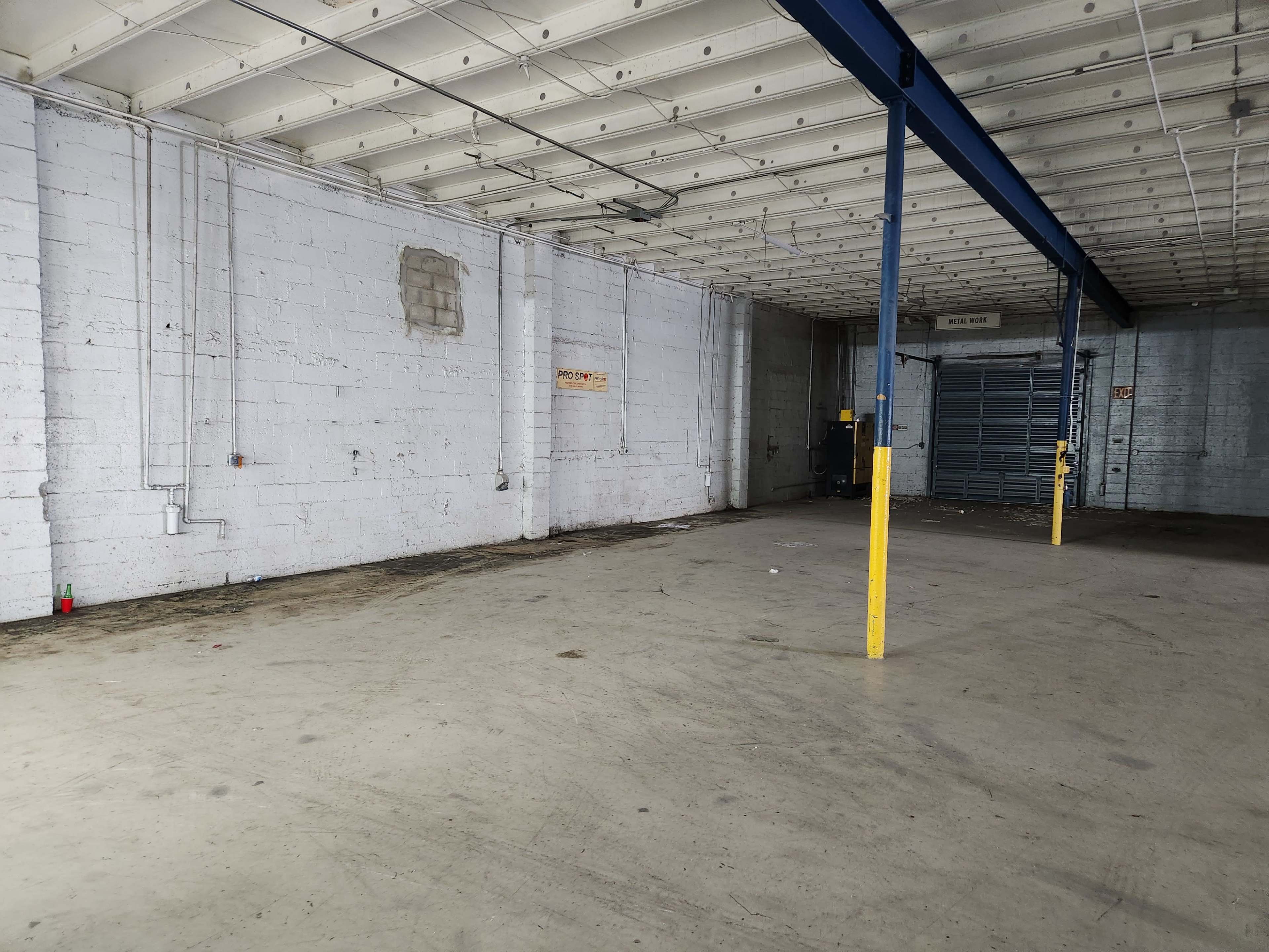 The image depicts a large, empty warehouse space with concrete flooring and exposed walls, featuring a few structural supports and minimal equipment along the sides.