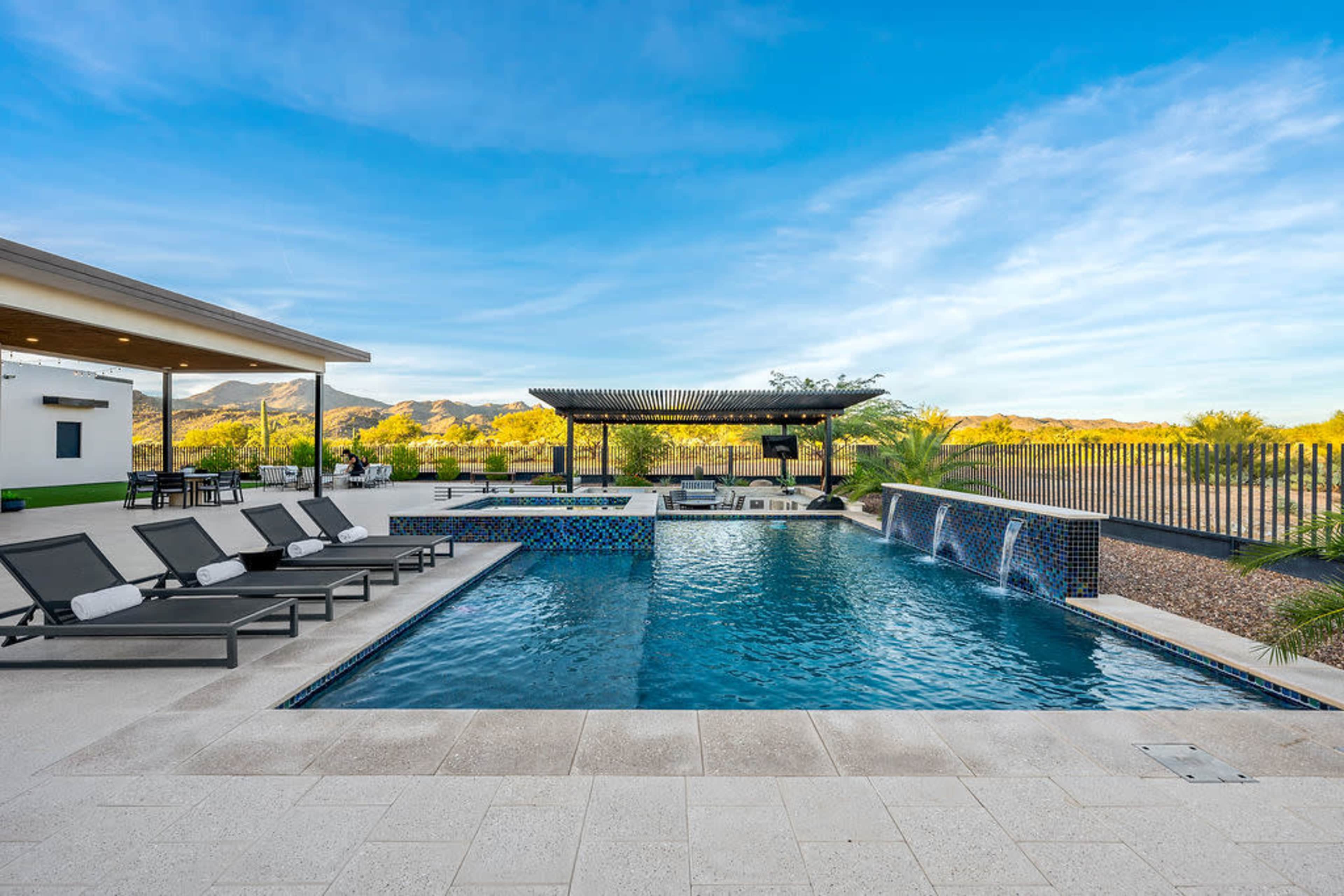 The image shows a modern outdoor swimming pool with lounge chairs, a shaded seating area, and a backdrop of mountains under a clear blue sky.